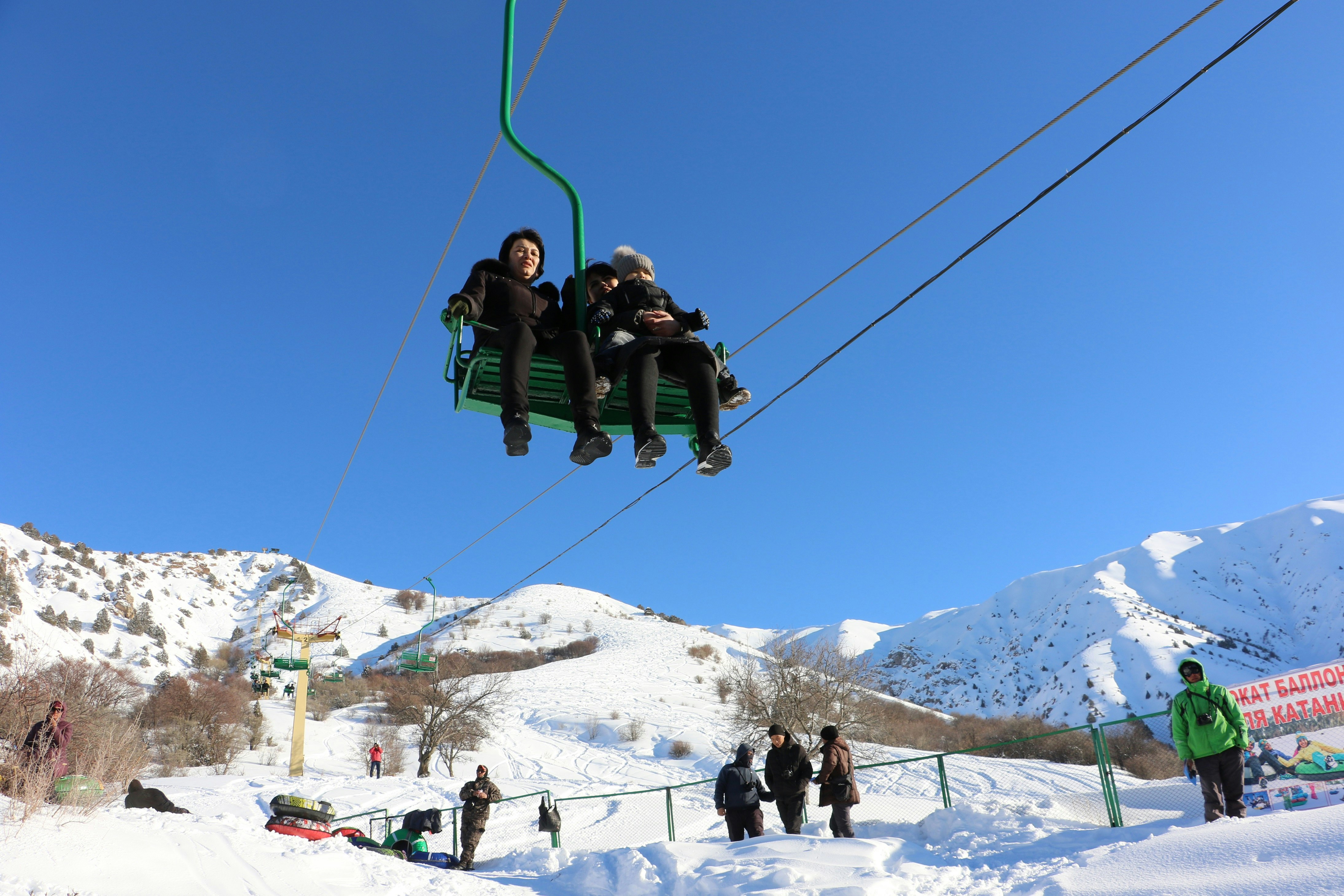 Three people (two adults and a child) ride in a chairlift at the Chimgan ski resort in Uzbekistan. The surrounding scenery is blanketed in snow and the sky is clear blue in the background.