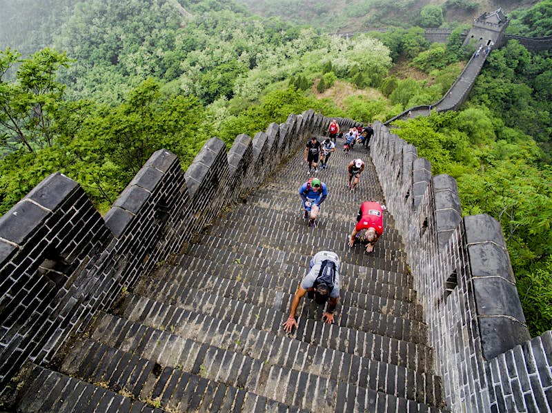 Competitors clamber up a steep section of steps on the Great Wall of China during the marathon; the wall descends behind them into the distance, with trees on each side of it.