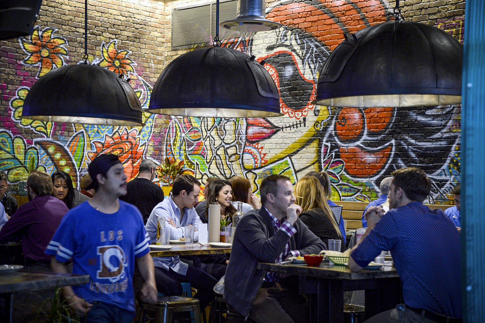 People enjoy tacos at tables beneath three umbrella-shaped lamps. The wall is brick and painted with a brightly colored mural depicted a person wearing sugar skull makeup. Denver, Colorado.