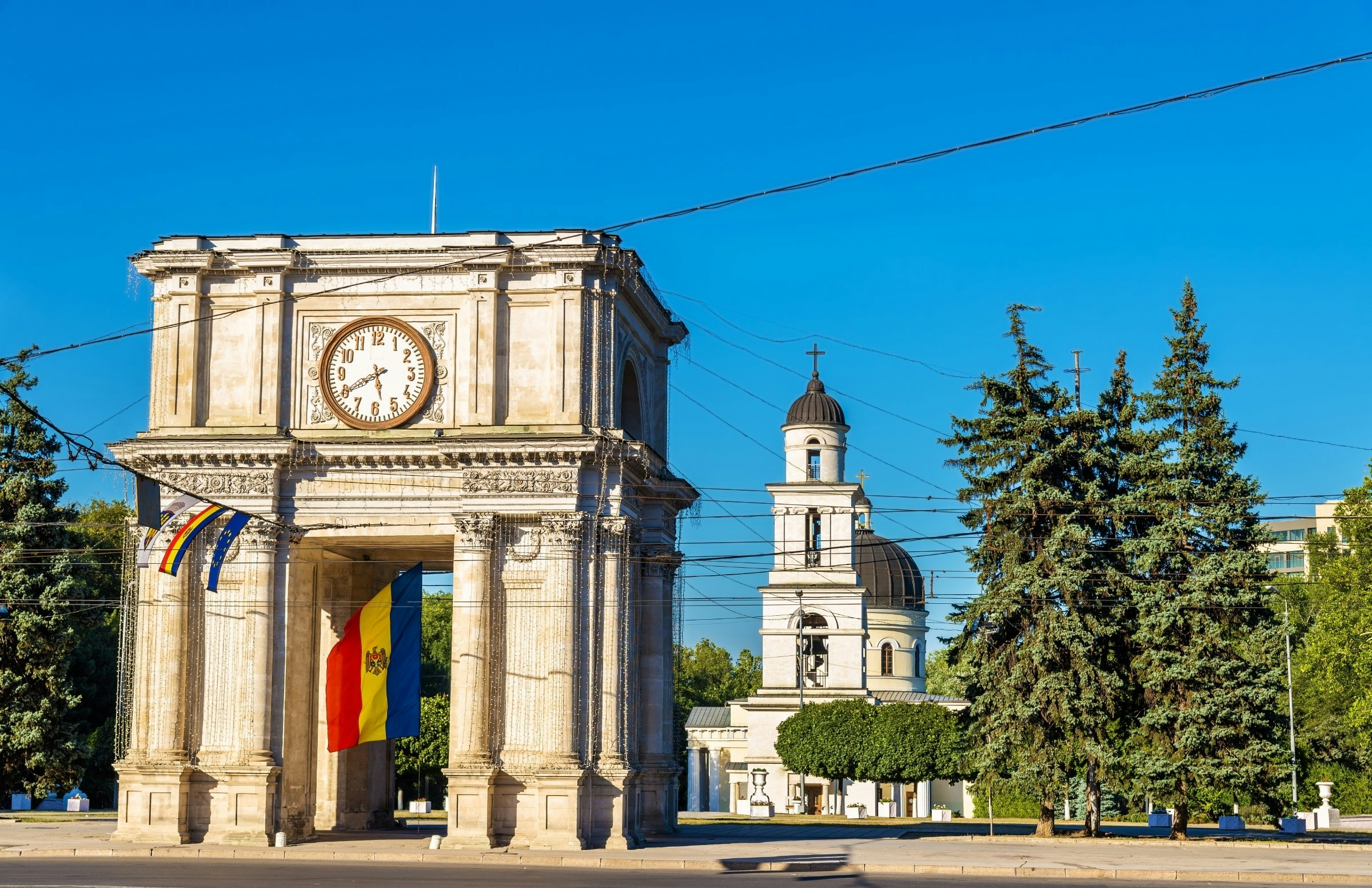 A large, white stone, freestanding arch in a city square with a church building behind it