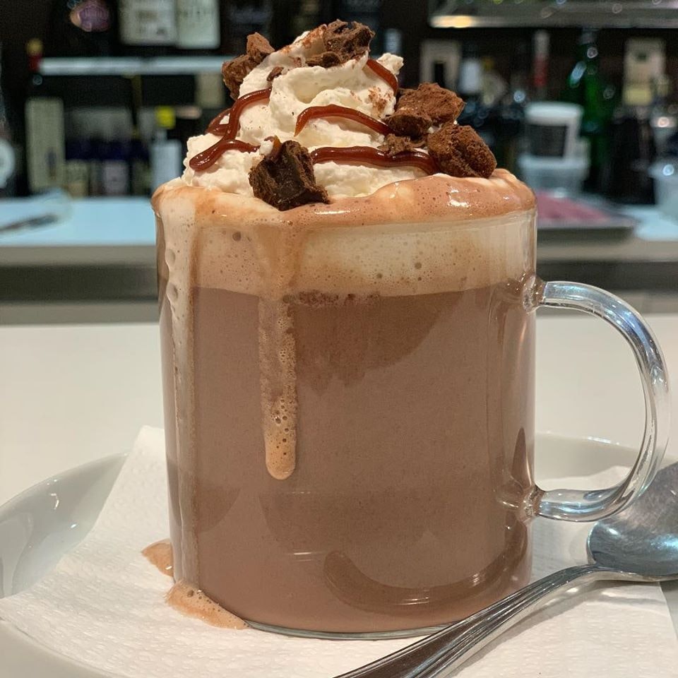 An overflowing glass mug of hot chocolate topped with whipped cream, chocolate sauce, and chocolate chunks