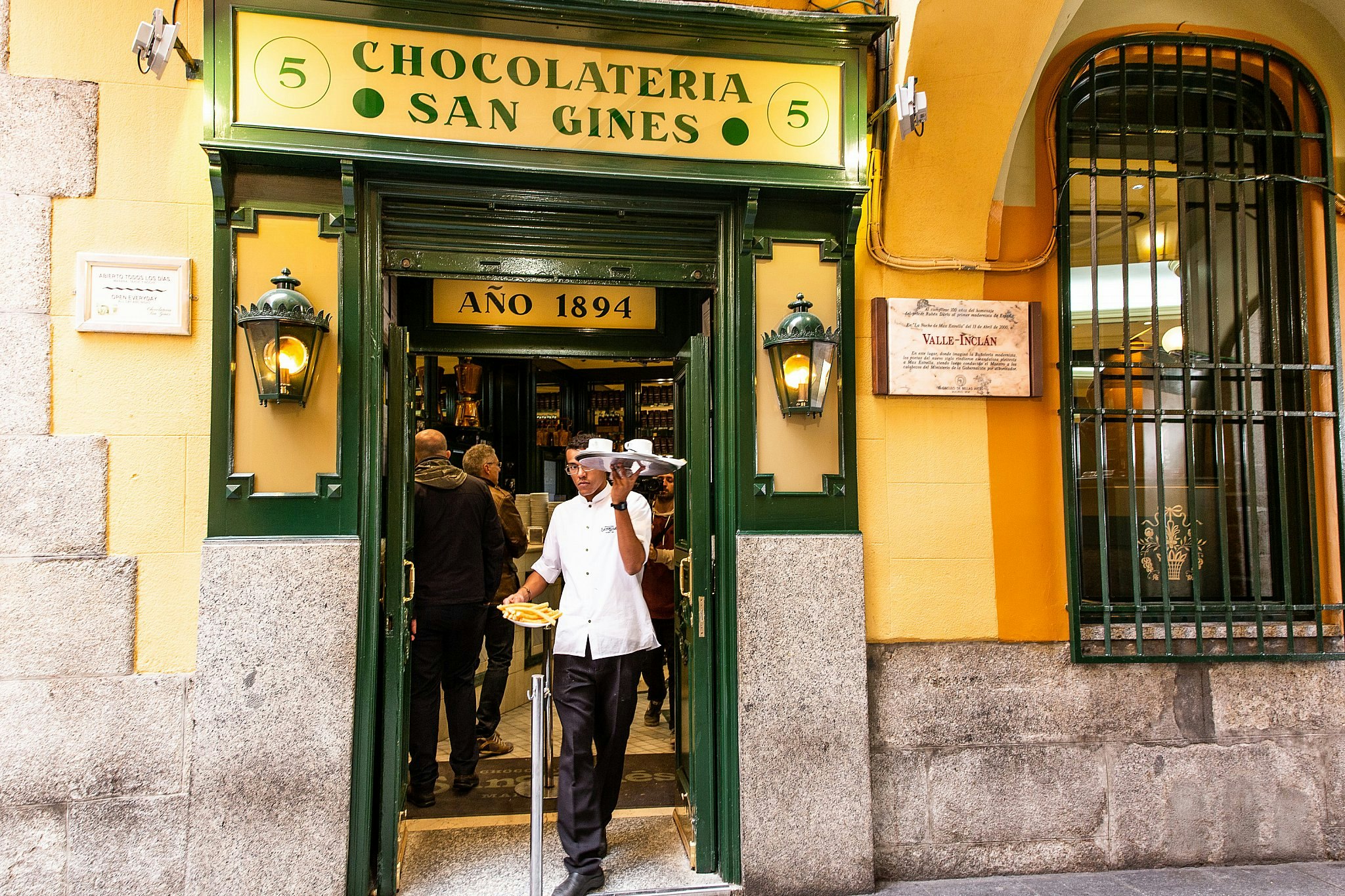 A waiter bearing plates of churros and chocolate emerges from Chocolatería San Ginés Madrid; the exterior is painted yellow with a green door frame and signs.