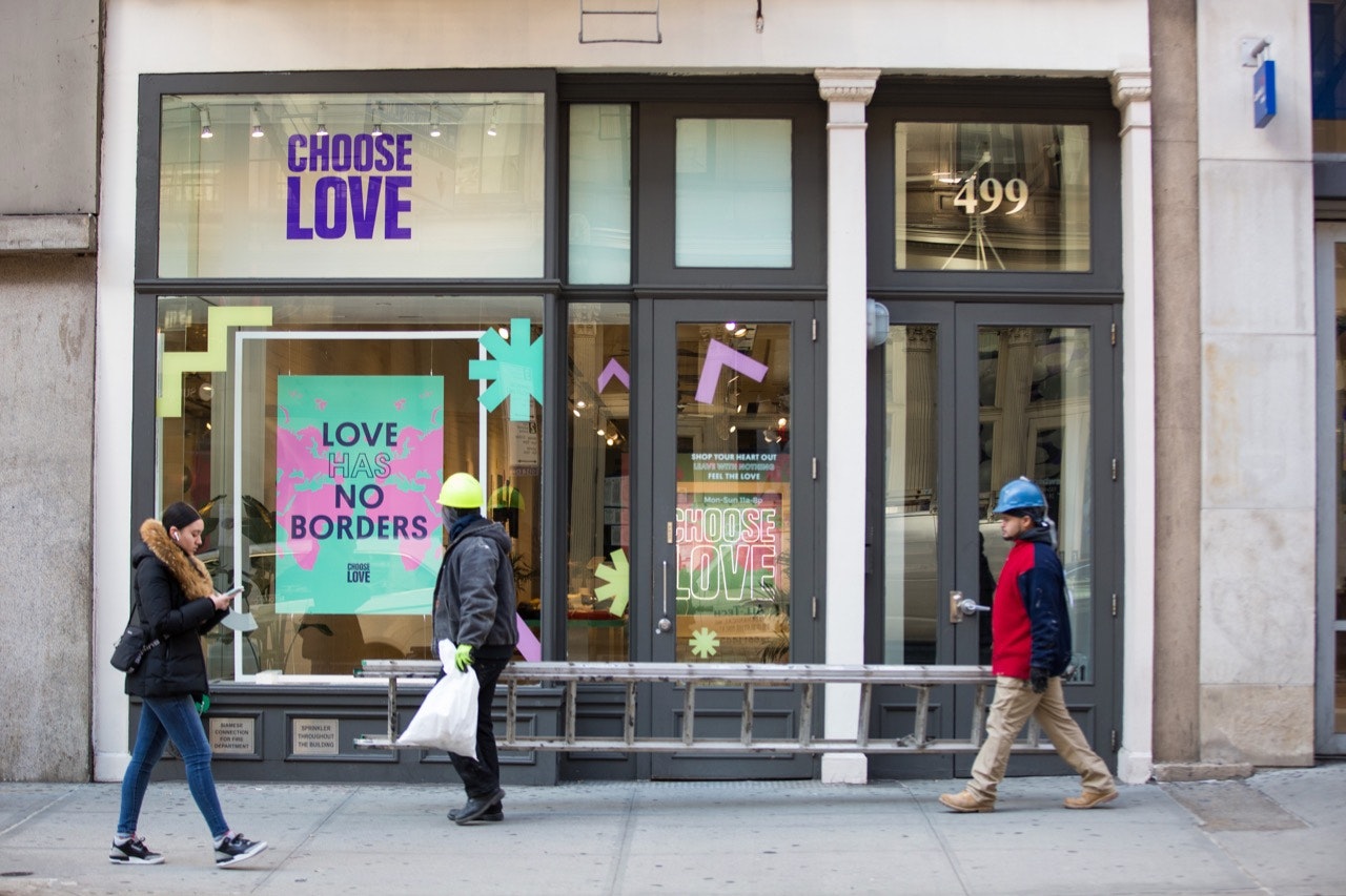 A person looking at their phone and two construction workers carrying a ladder walk by the NYC Choose Love store in Soho