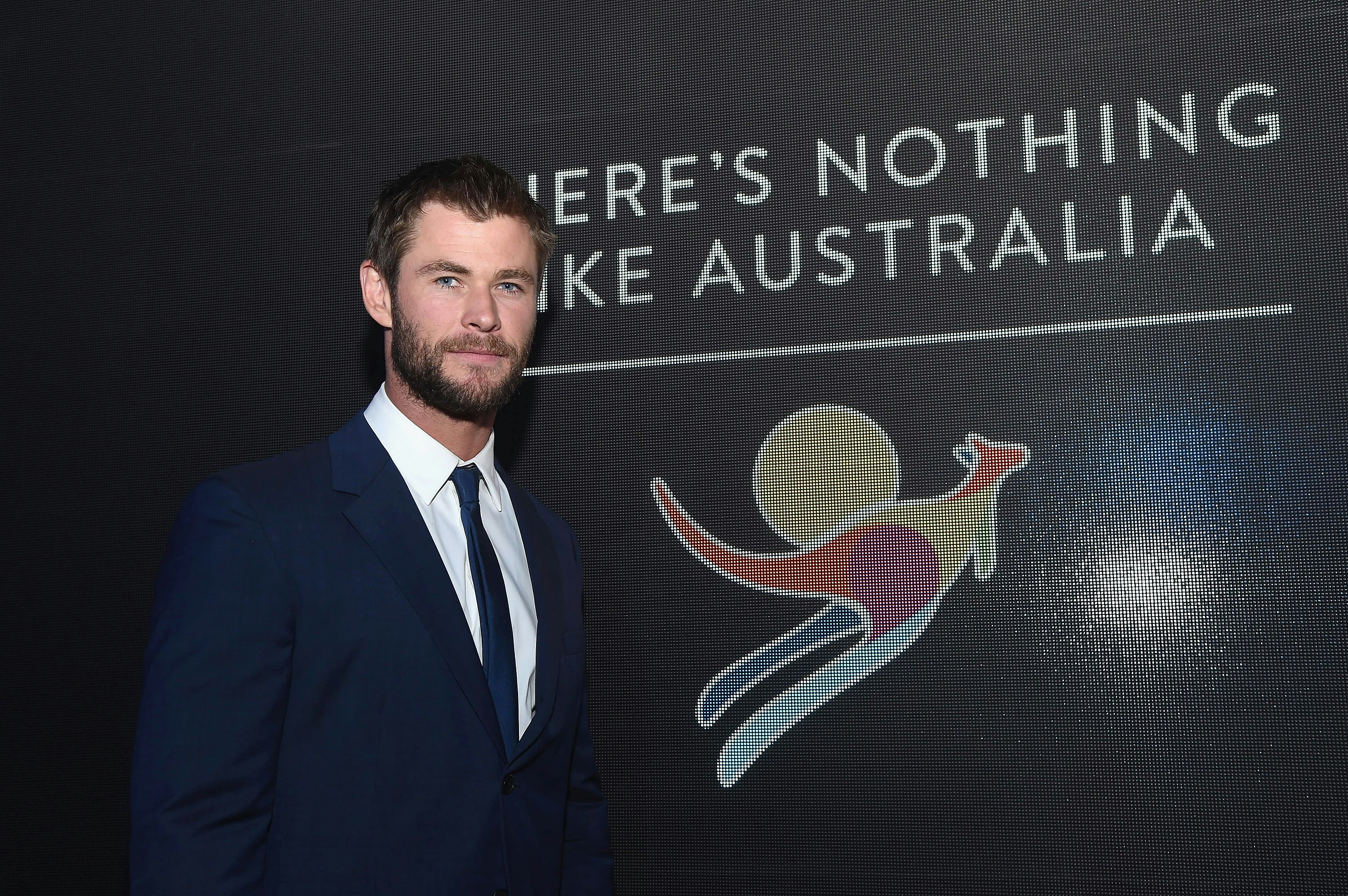 Chris Hemsworth stands in front of a Tourism Australia poster. 