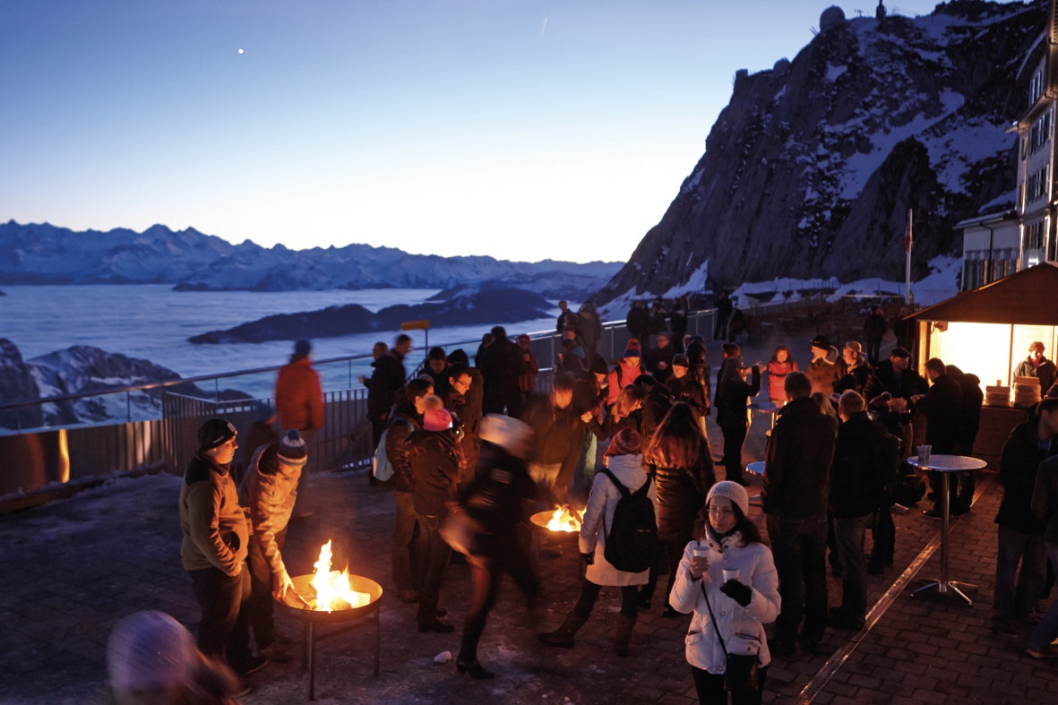 People gathered on a platform on the side of a rugged, snow-covered Mt Pilatus, Switzerland. There are fires and stalls selling food and drink as part of Christkindlimärt