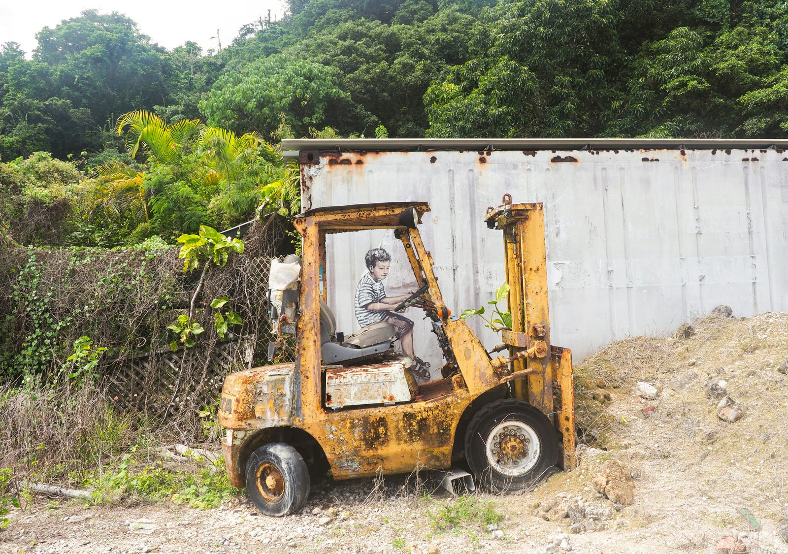 A forlorn forklift is parked next to an old shipping container; an image of a boy has been painted on the container to look as if he is driving the forklift