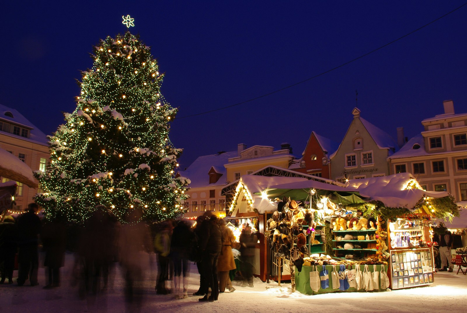 Christmas market in Tallinn, Estonia, at night. There is a huge decorated Christmas tree among the wooden stalls selling Christmas trinkets. There are blurred people walking through the market.