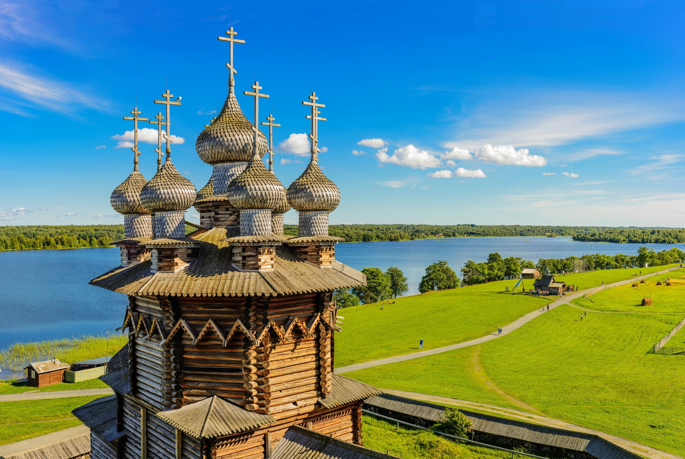 A multi-domed wooden tower, with each dome topped with a cross, set within a grassy field on the edge of a lake