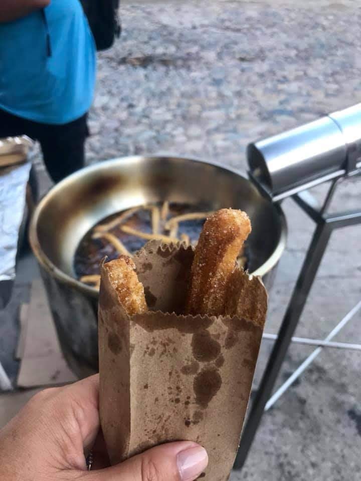 A hand holds an oiled-stained brown paper bag filled with two fresh churros, dusted with sugar, on a street in Puerto Vallarta