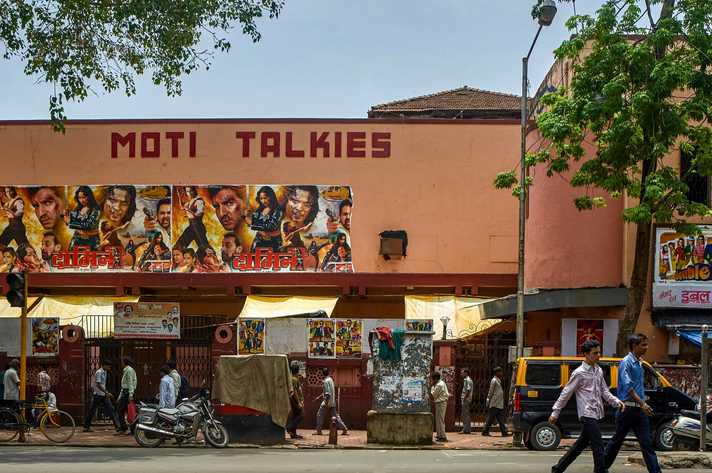 The exterior of the Moti Talkies, one of many cinemas in Mumbai. The building is large, and painted in a terracotta colour, with many people walking past in the street.