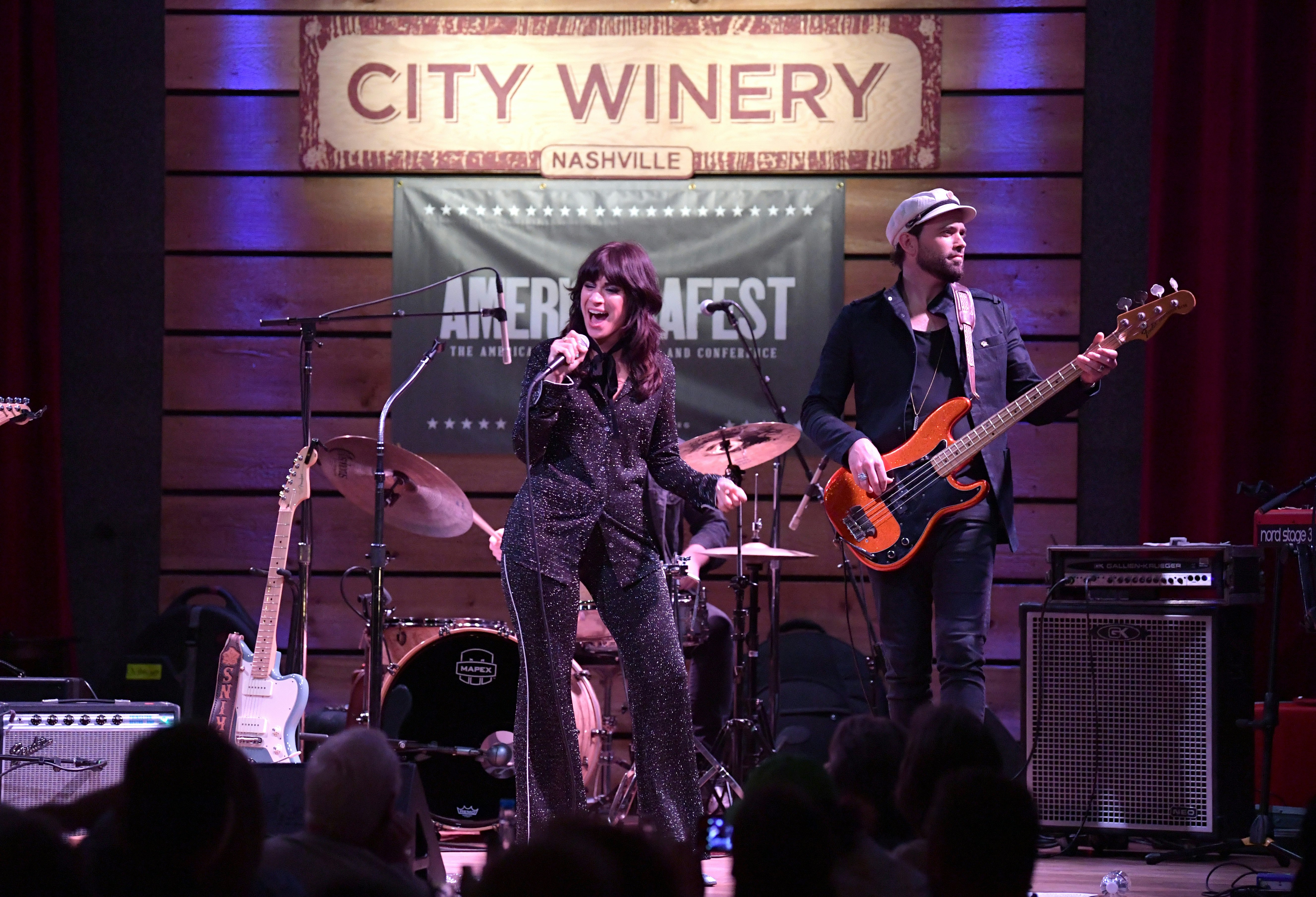 En kvinna i en glittrig kostym sjunger i en mikrofon på en scen bredvid en man som spelar orange gitarr på City Winery i Nashville.