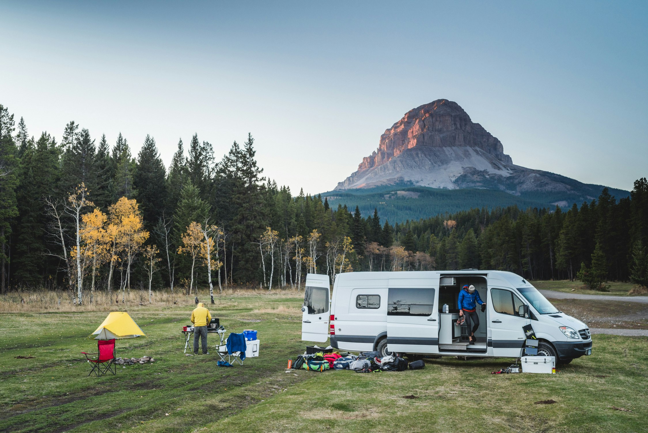 a white campervan parked in a grassy field surrounded by trees and mountains and people setting up camp