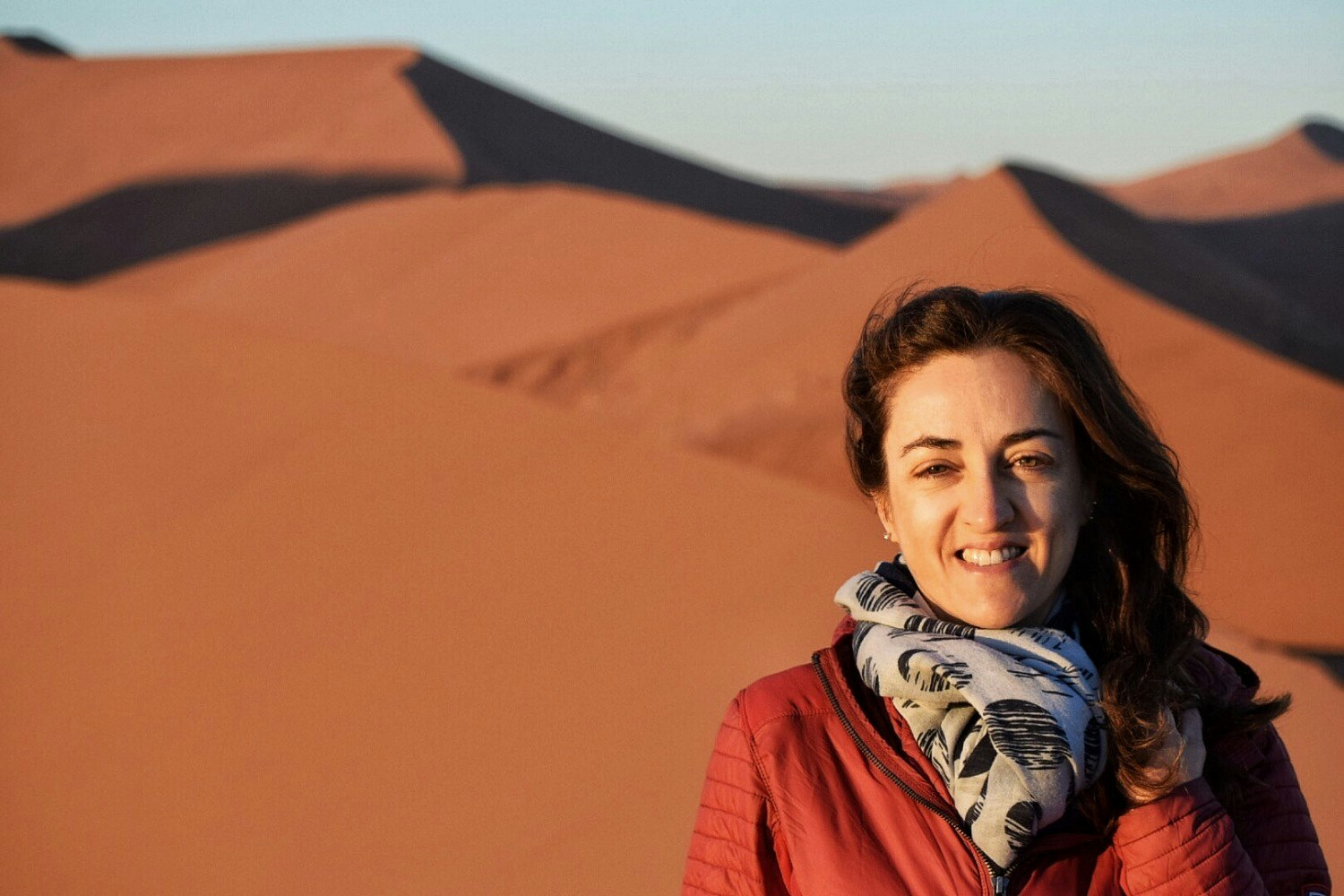 Claudia poses in front of steep sand dunes in a red jacket