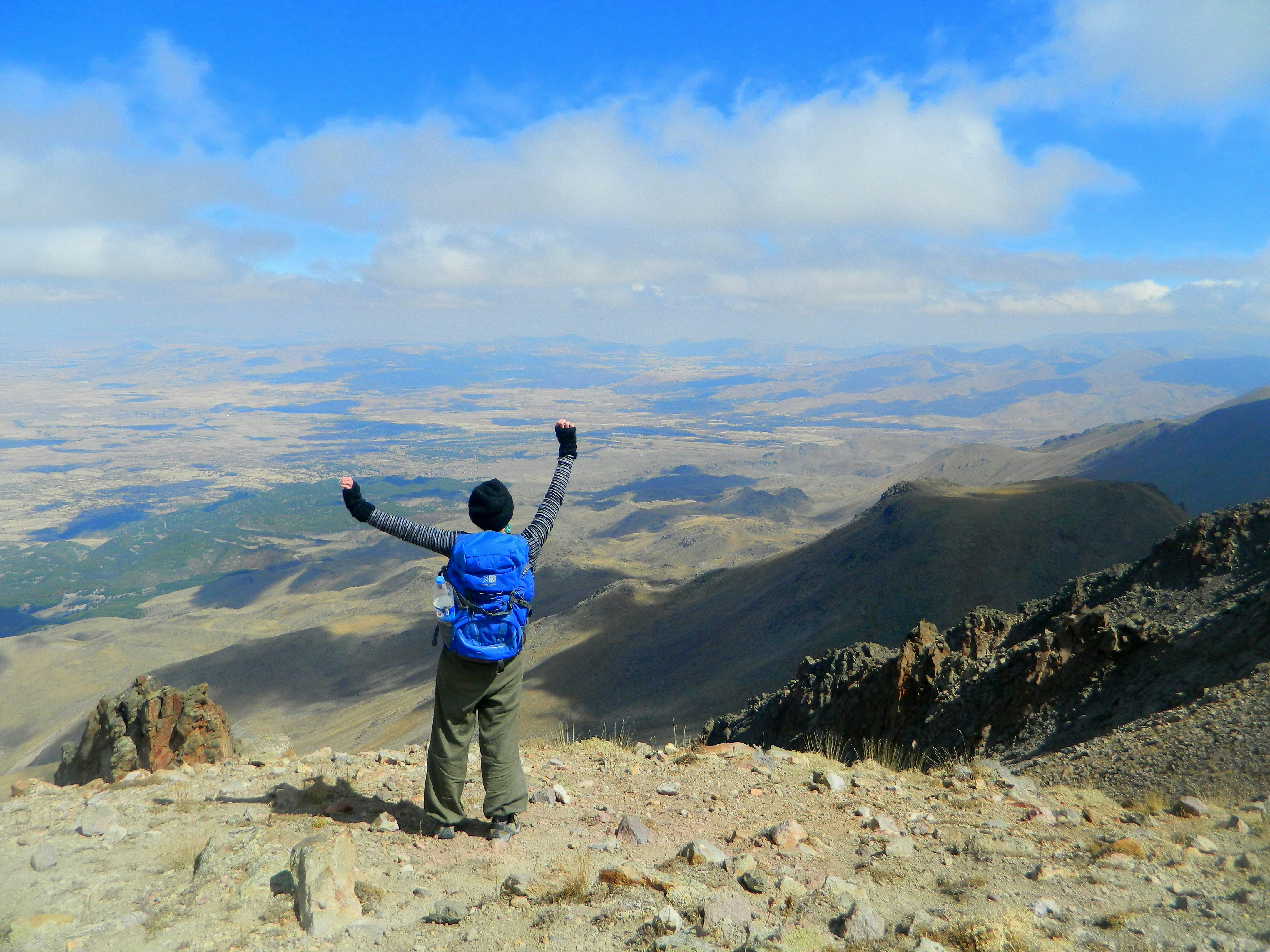Write Jess Lee climbing Mt Hasan, Turkey; she faces away from the camera waving her arms aloft, staring over a barren yet beautiful landscape.