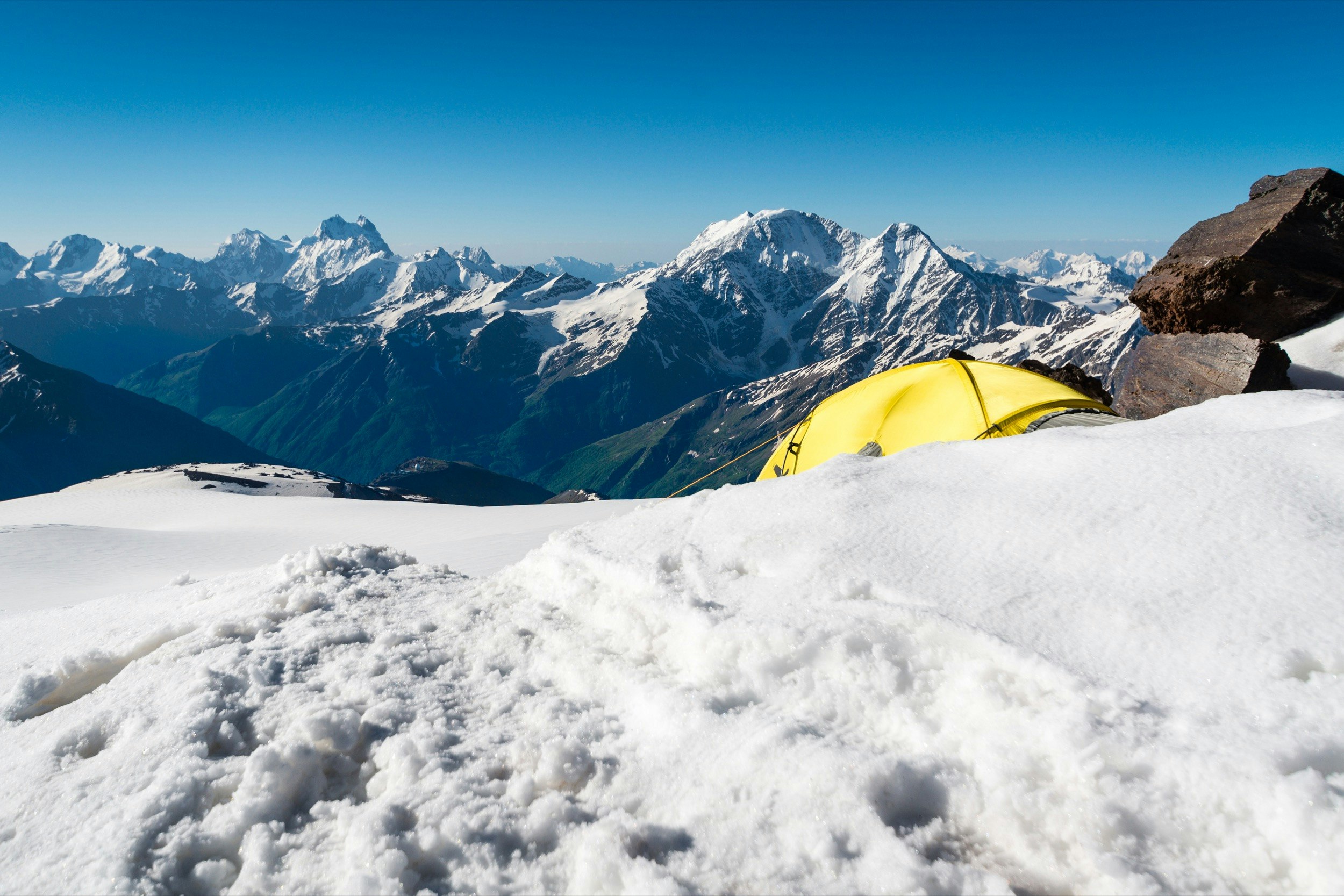 A yellow tent is partially obscured by a snow bank near the top of a very tall mountain in Russia; Climbing fourteeners for beginners