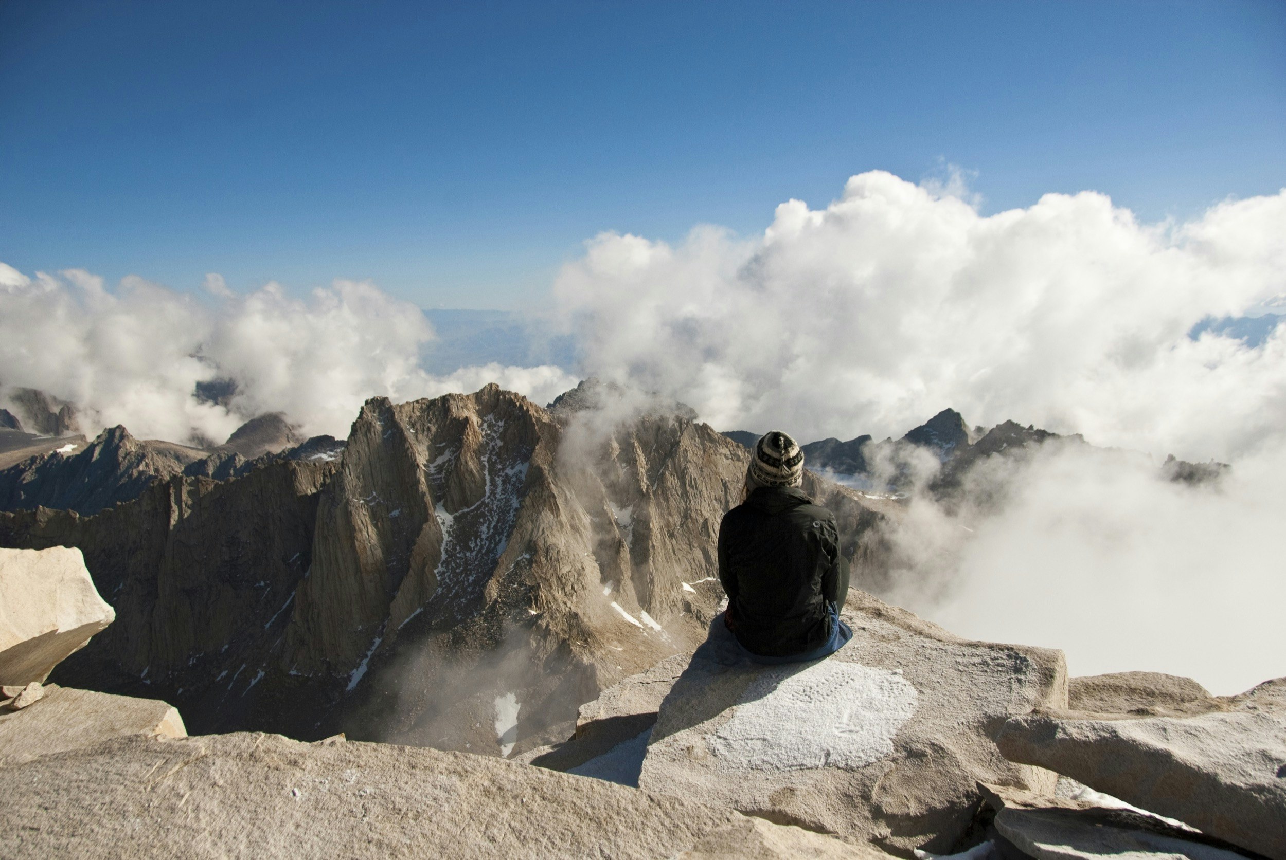A woman sits at the summit of a mountain looking down at the clouds mixing around the surrounding peaks; Mountain climbing fourteeners