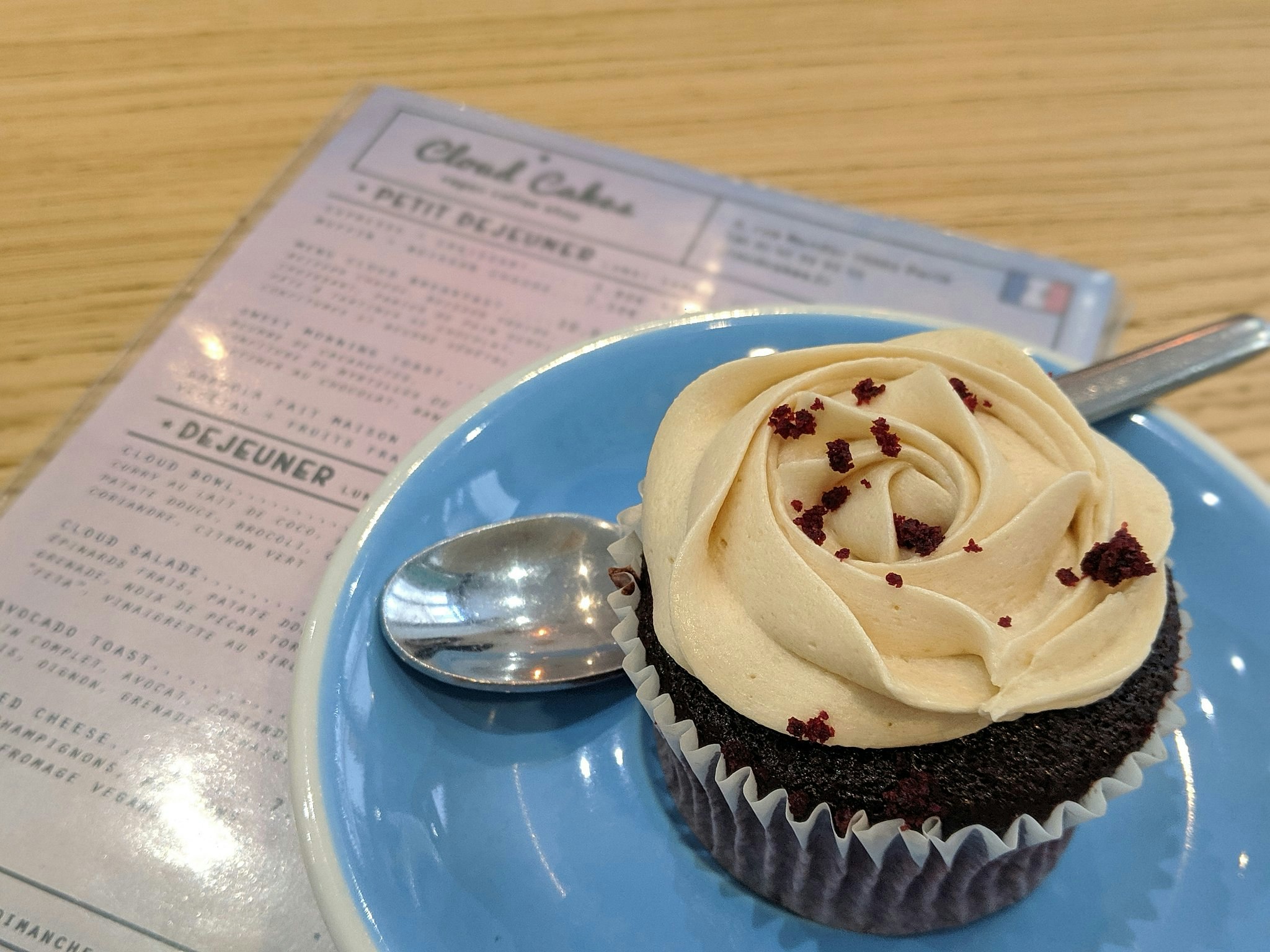 A chocolate-coloured muffin with cream frosting on a blue plate with a silver spoon next to a menu.