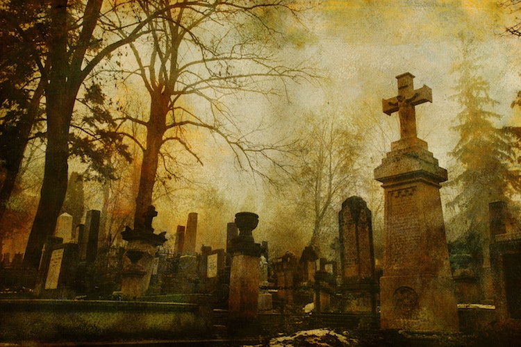 A sepia-toned photo of old cemetery grave stones and bare tree branches; haunted places world 