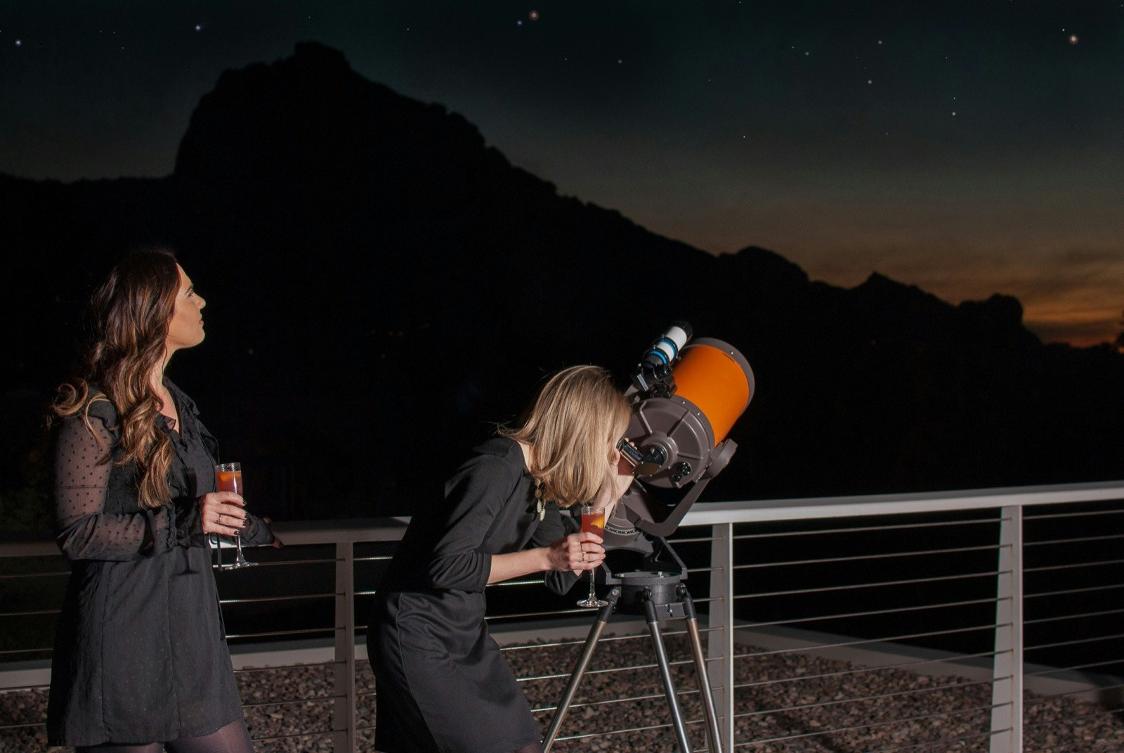 Two women hold cocktails while one looks through a telescope at a night sky