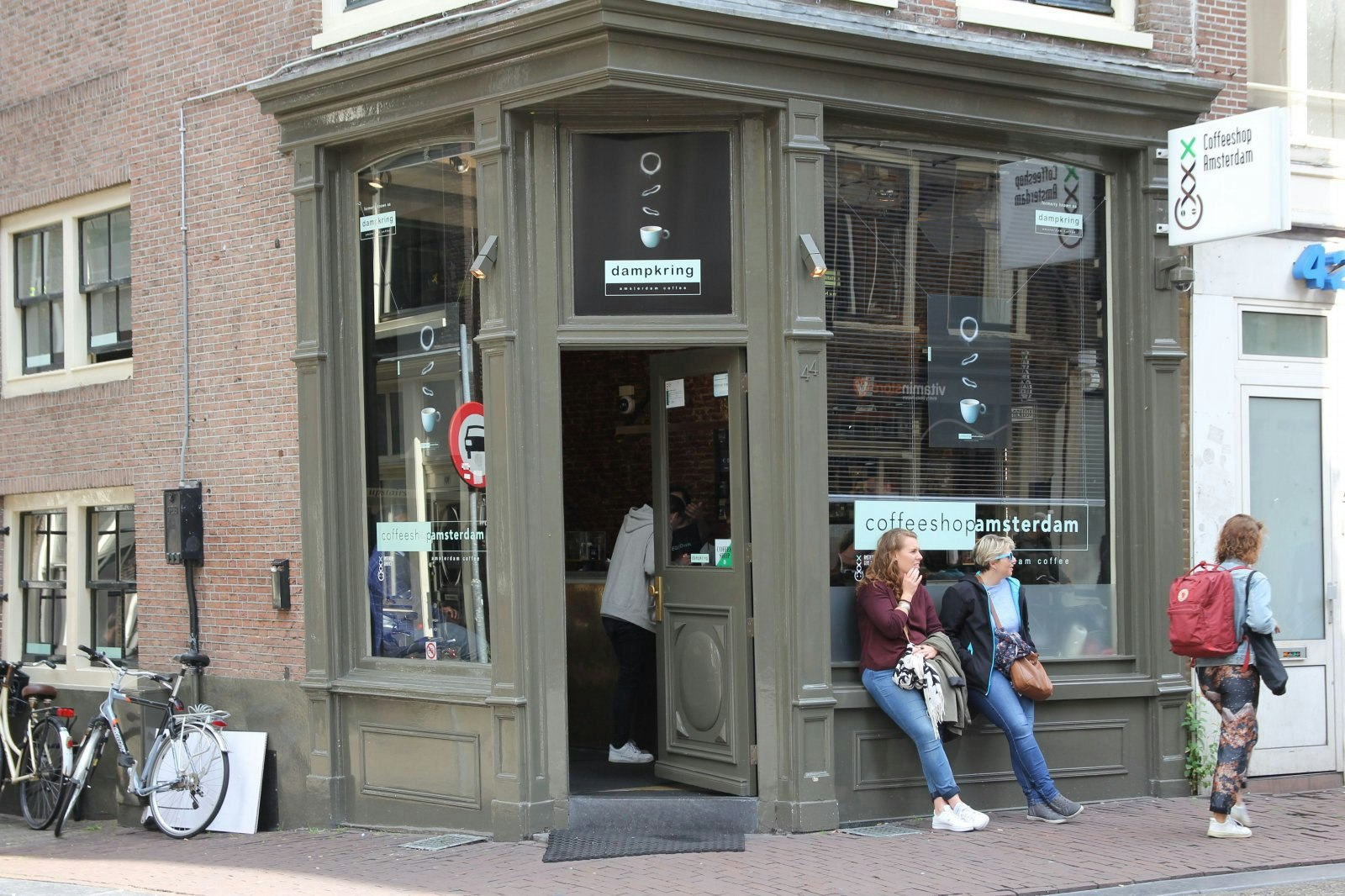 Exteriören på Coffeeshop Amsterdam som är målad i mörk khaki.  De stora fönstren har butikens logotyper och skyms av persienner.  Två kvinnor lutar sig mot fönsterkanten utanför butiken. 