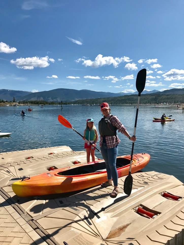 A woman and her daughter pose with kayaks by the side of a lake 
