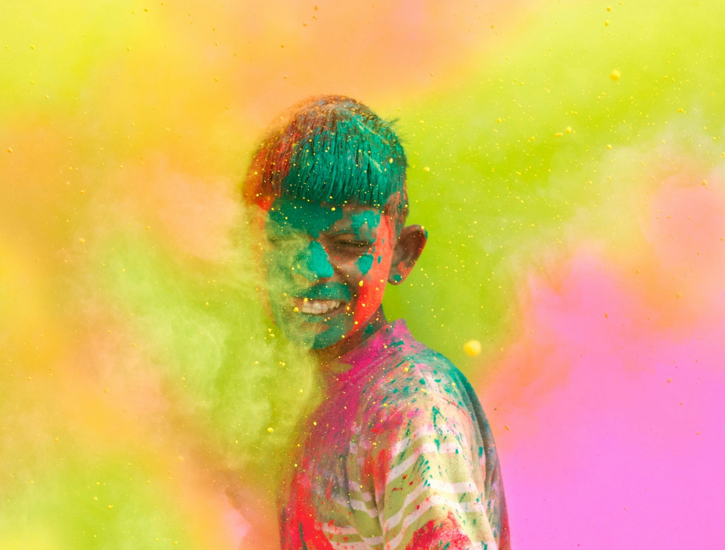 A young boy is pelted with handfuls of colourful powder, making for a colourful scene. The boy grins in the centre of the shot.