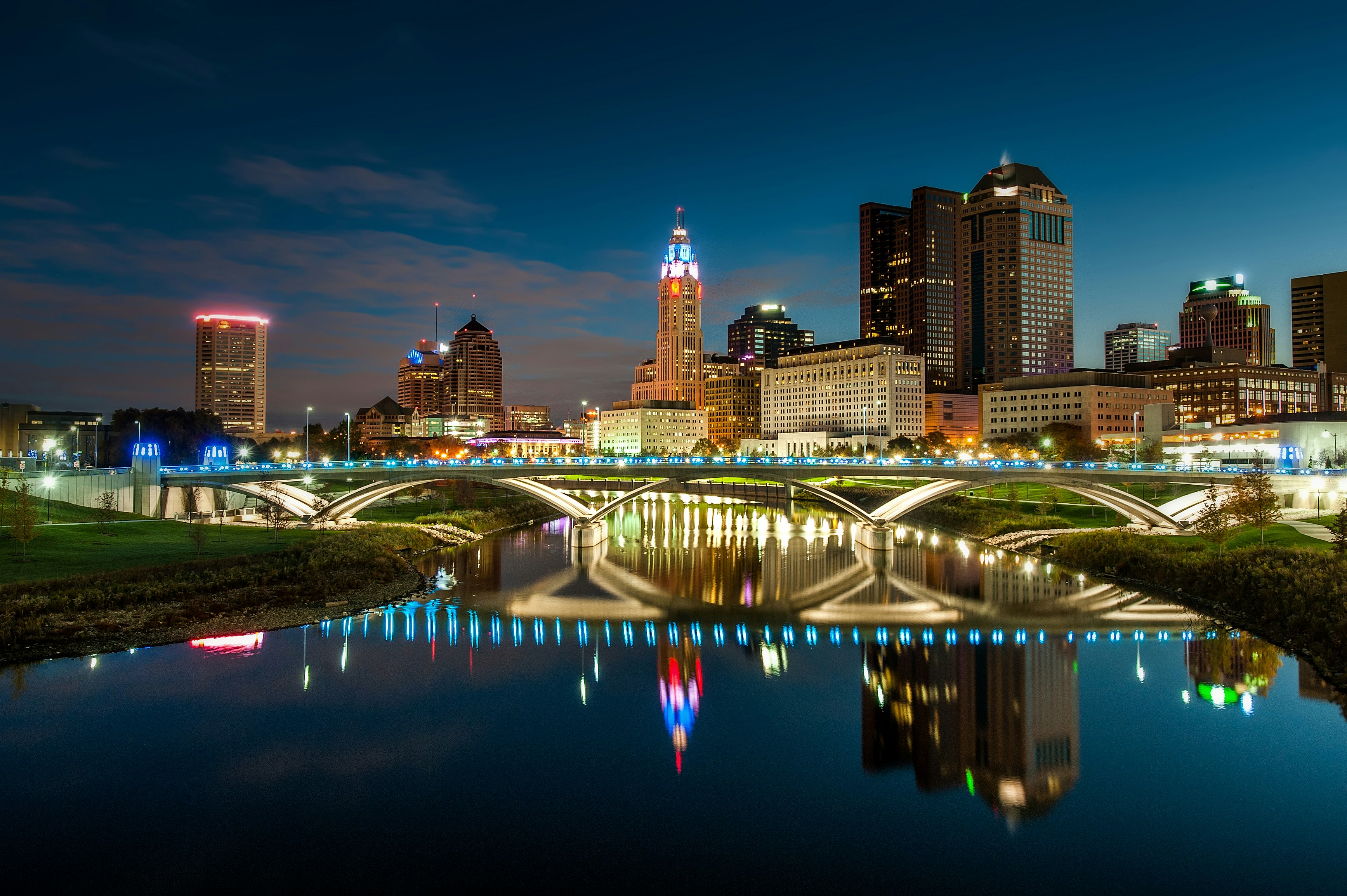 The bright city lights of downtown Columbus is mirrored in a body of water under flowing under a stone bridge