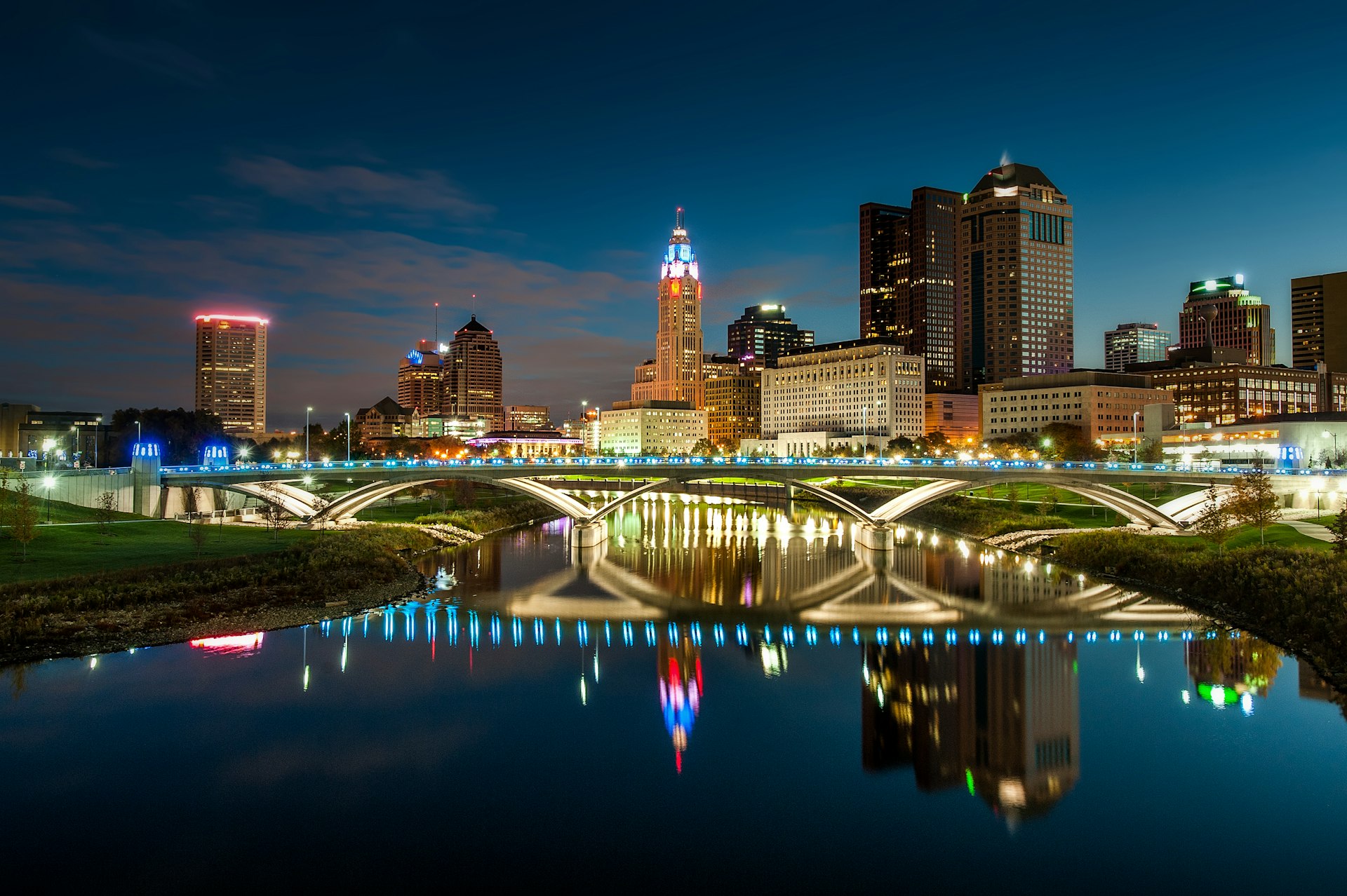 The bright city lights of downtown Columbus is mirrored in a body of water under flowing under a stone bridge; Columbus vs AnnArbor 