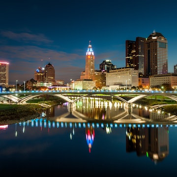 The bright city lights of downtown Columbus is mirrored in a body of water under flowing under a stone bridge
