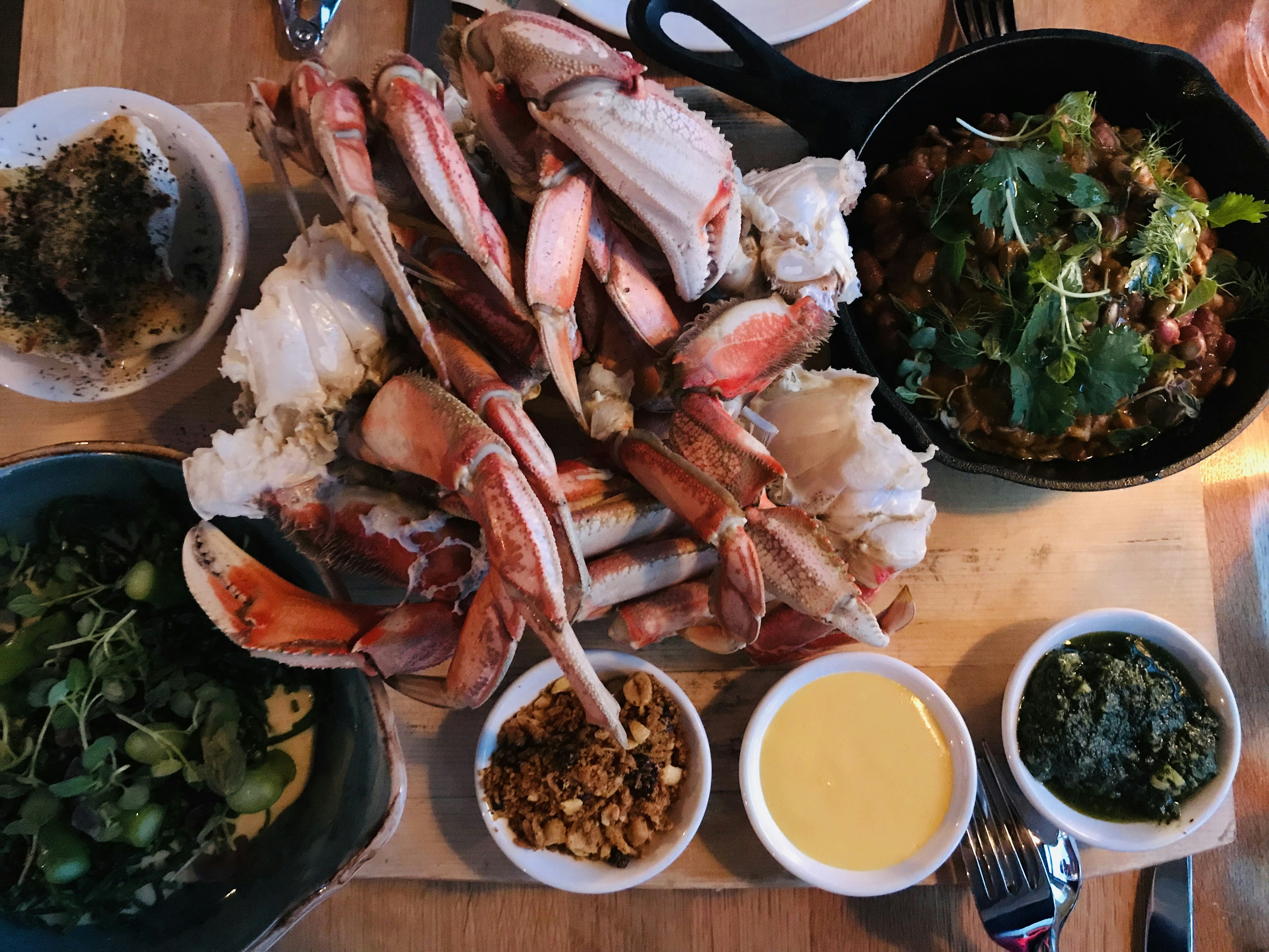 A blonde wooden table is laden with dishes at 1909 Kitchen. In the center is a huge pile of pink and white crab legs. To the lower left is a deep blue bowl with foraged micro-greens. Three small bowls are filled with peanuts and sauces. A small cast iron skillet in the top right is full of a brown dish garnished with parsley and more bright greens.
