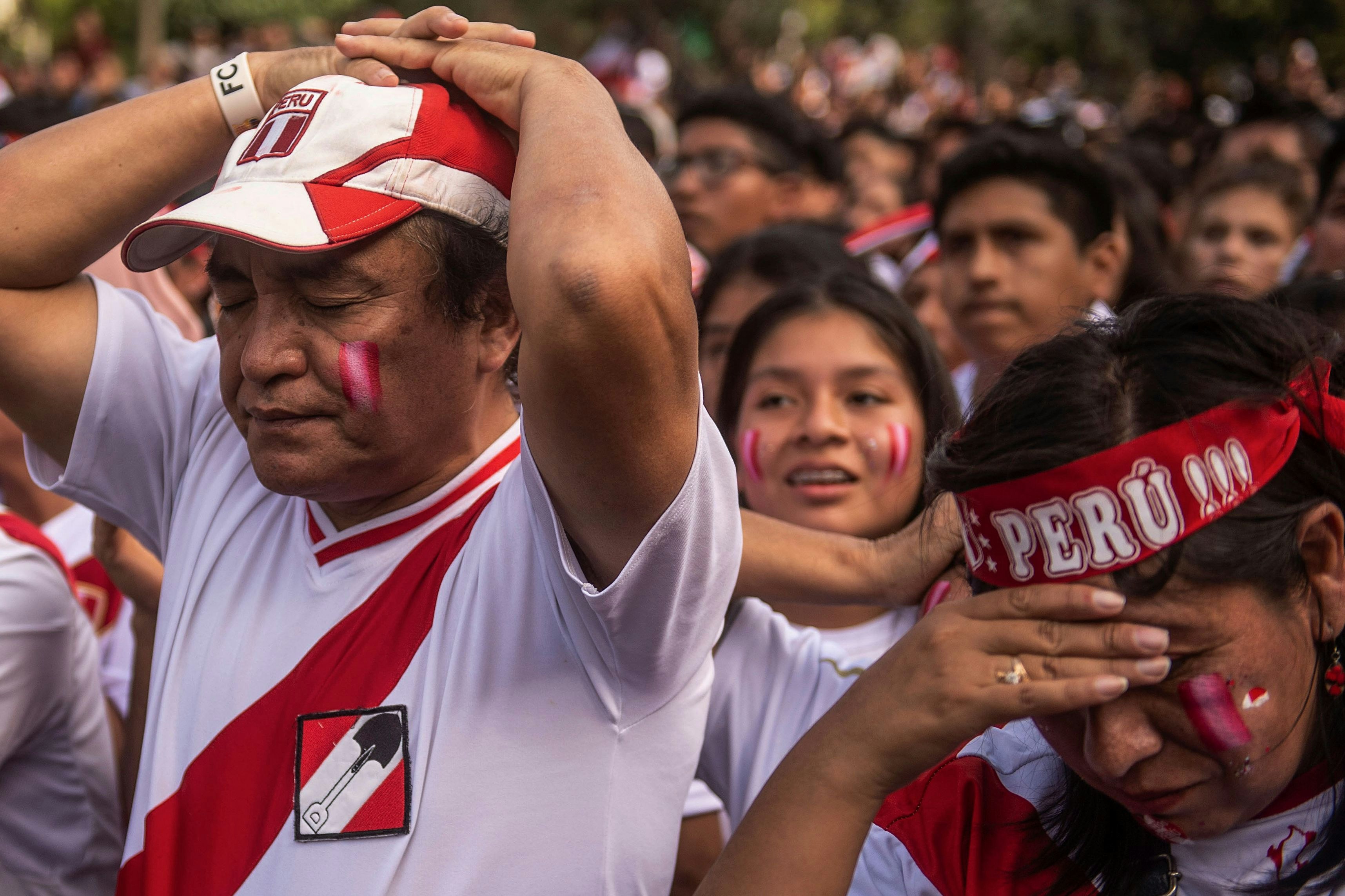 A man with his hands resting on the top of his head with his eyes close stands next to a woman wearing a "Peru" headband who has her hand resting on her forehead. They are surrounded by a large group of sad Peru fans after a soccer loss. 