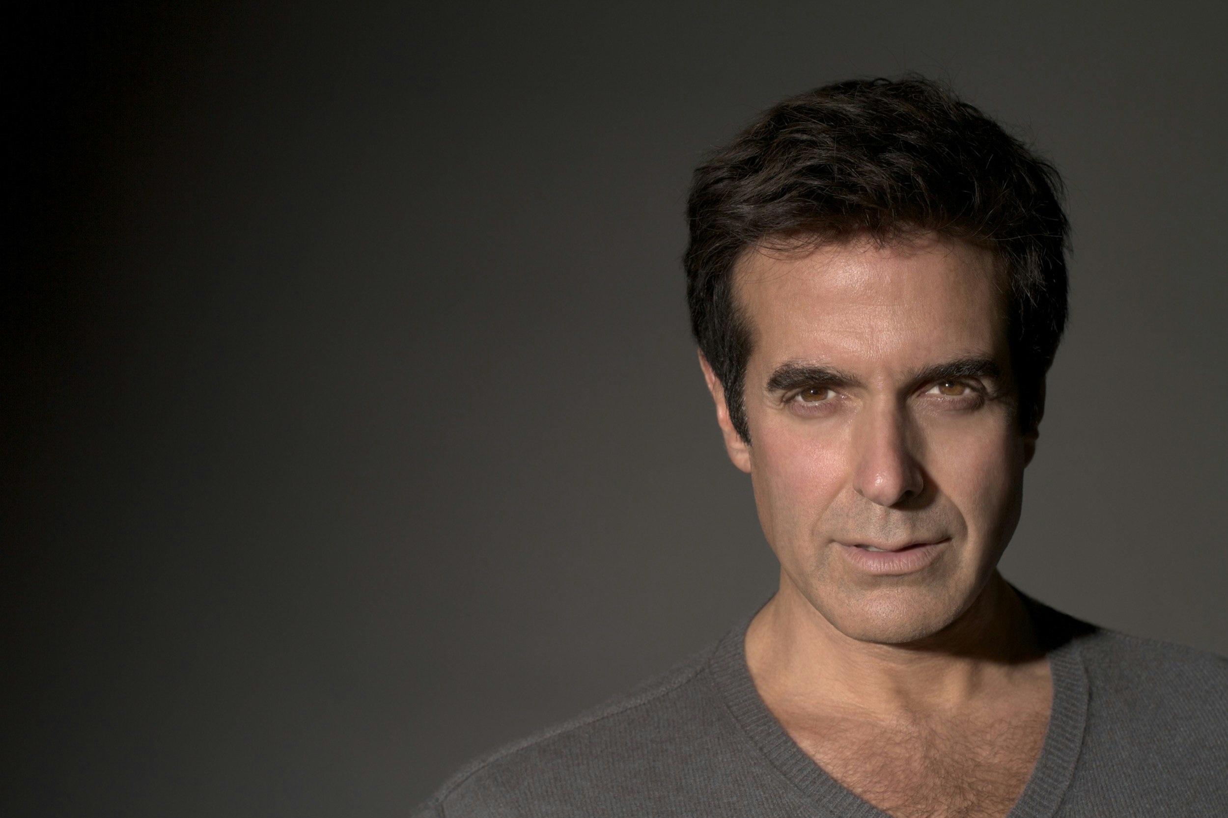 Magician David Copperfield wears a gray t-shirt on a darker gray background as his light brown eyes shine in a band of light