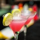 A pair of martini glasses filled with the bright pink cosmopolitans sit on a wooden bar. There are wedges of lemon on the rim.