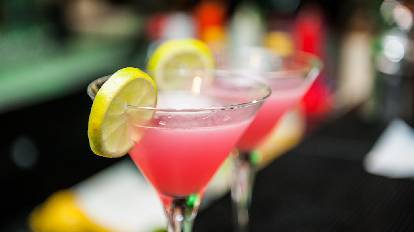 A pair of martini glasses filled with the bright pink cosmopolitans sit on a wooden bar. There are wedges of lemon on the rim.