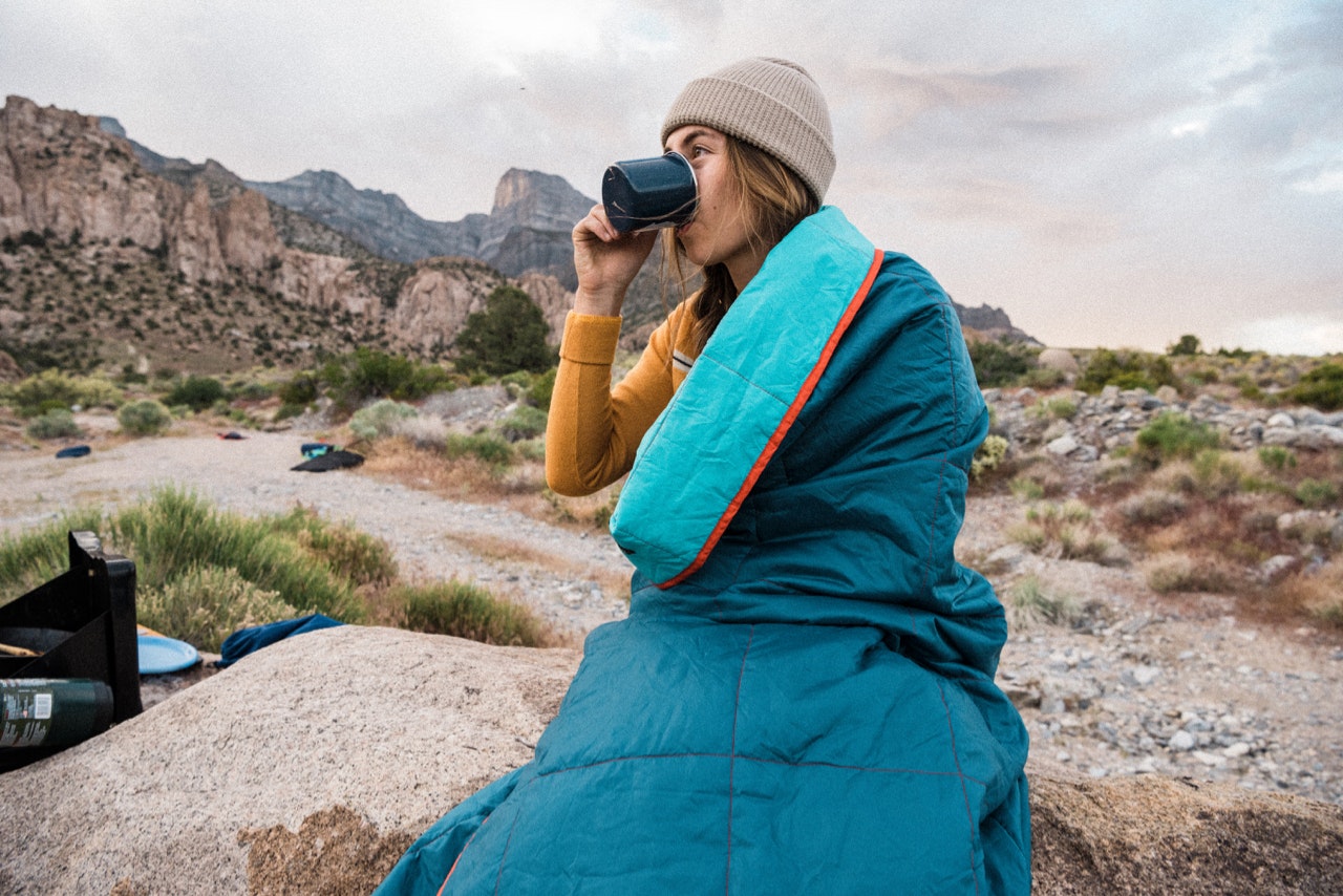 Woman in a yellow sweater and a beige hat at a campsite, wrapped in a blanket and drinking from a mug