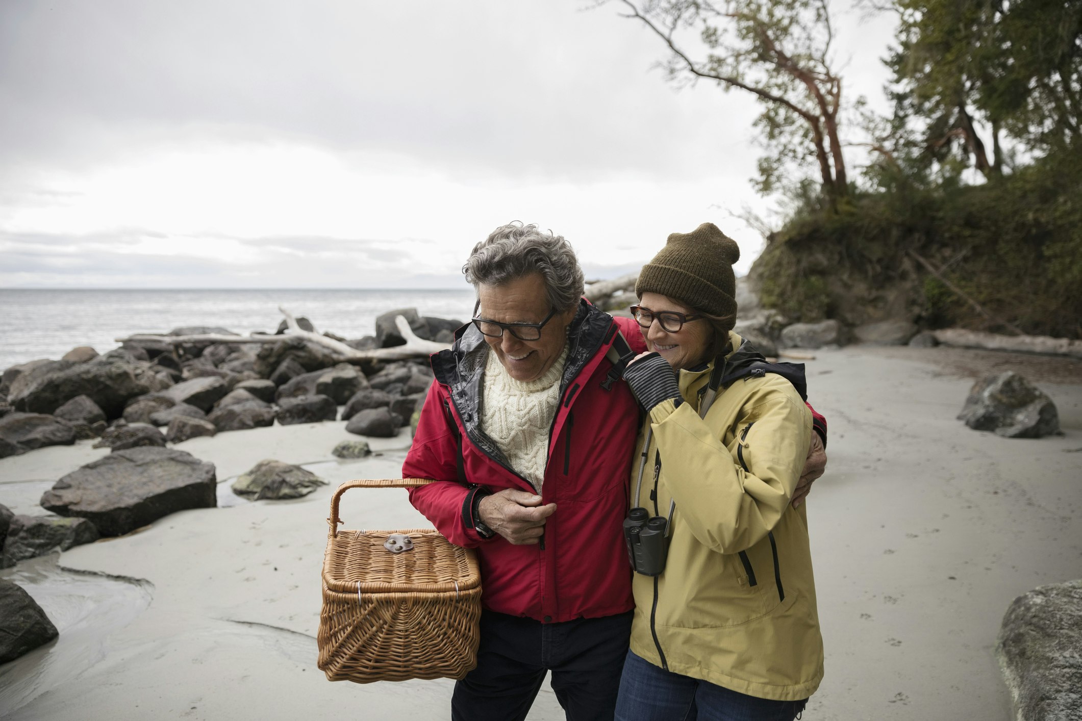 A couple laughs while carrying a picnic basket on the beach, dressed for a cold, drizzly day