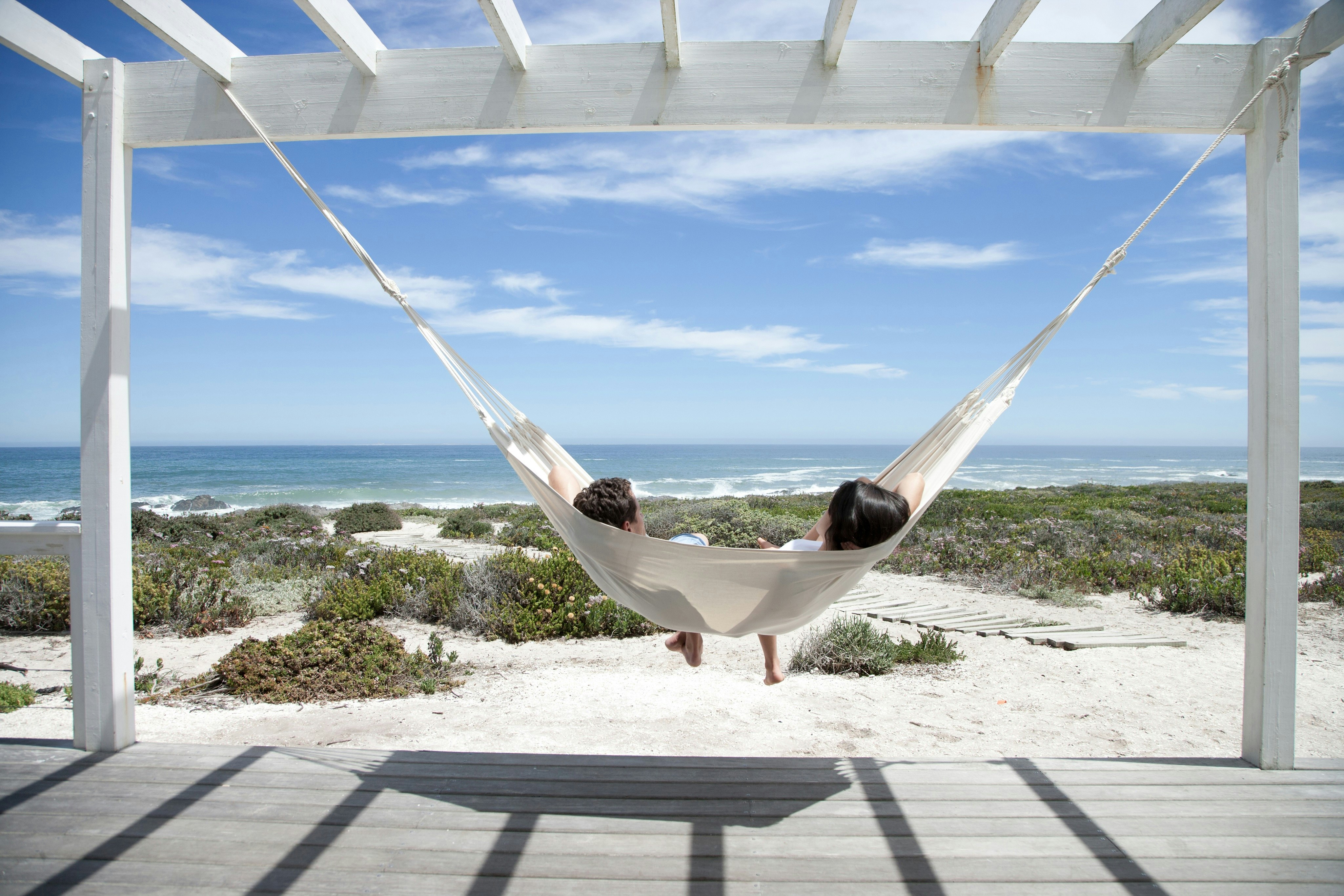 Two people doze serenely in a hammock facing a white beach and blue sea.