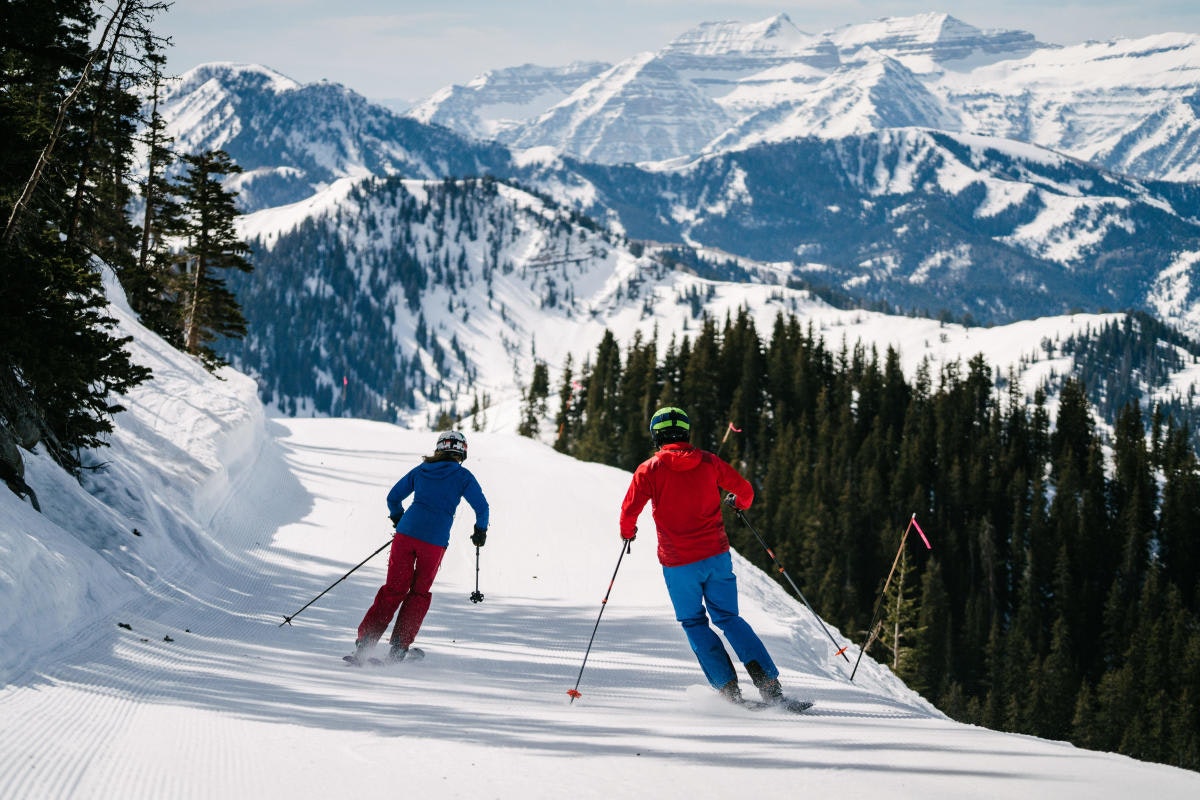 Two skiiers, one in red pants and a blue top and the other in blue pants and a red top, are viewed from behind as they ski down a white, unblemished slope with denim blue peaks capped with snow in the background.