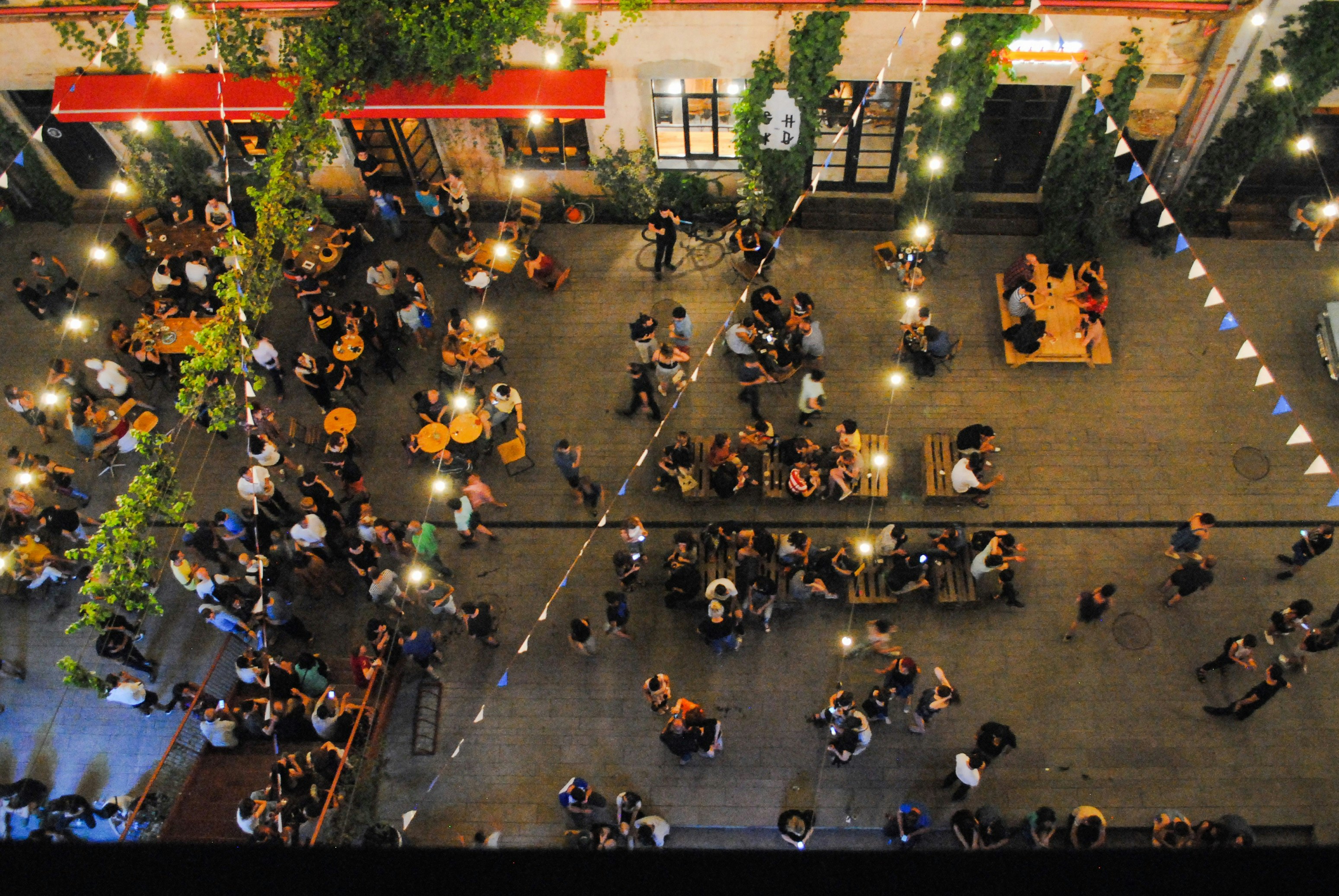 A shot downwards into a busy courtyard, where people are sitting at tables and standing in groups talking.