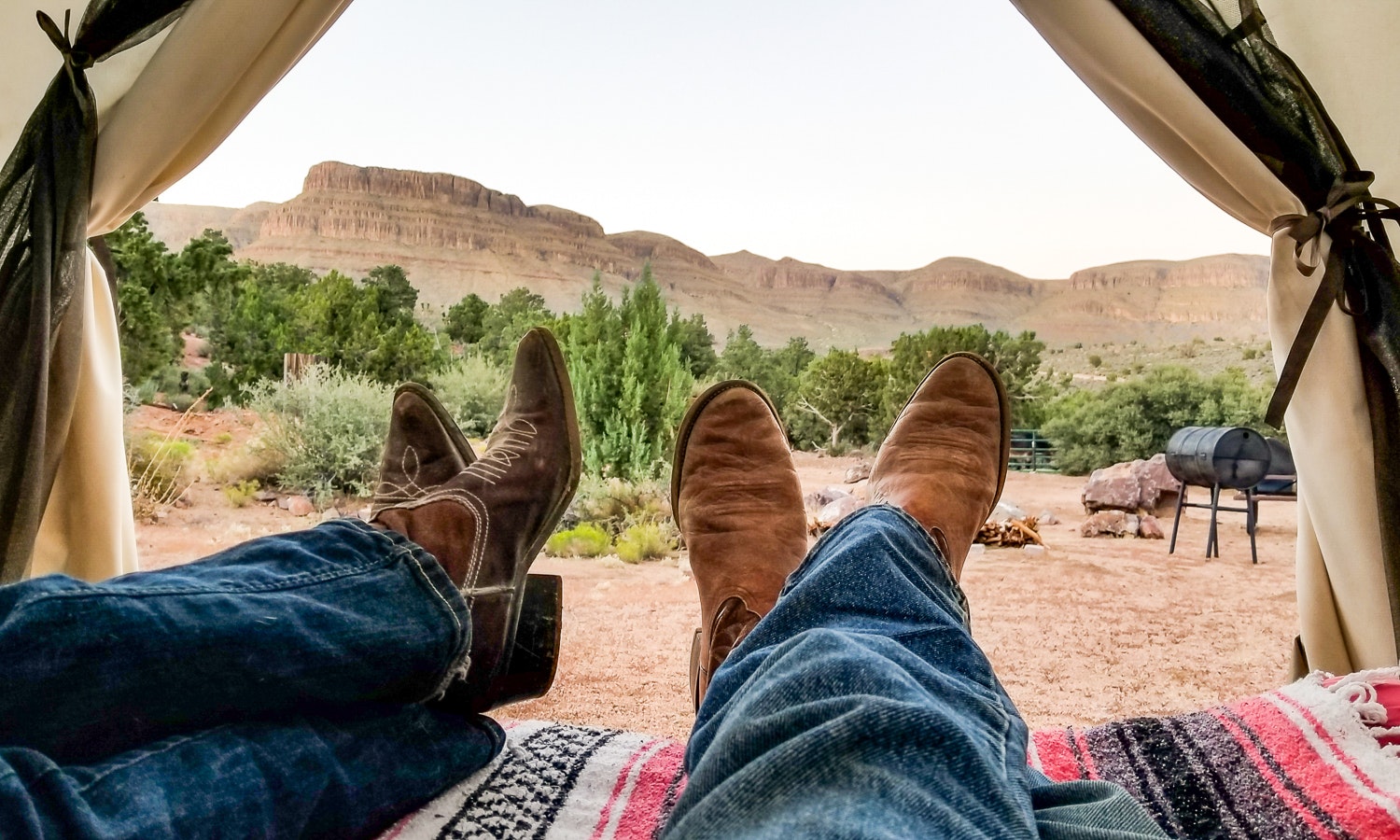 Two sets of cowboy-booted legs crossed under the opening of a tent, with a western view outside