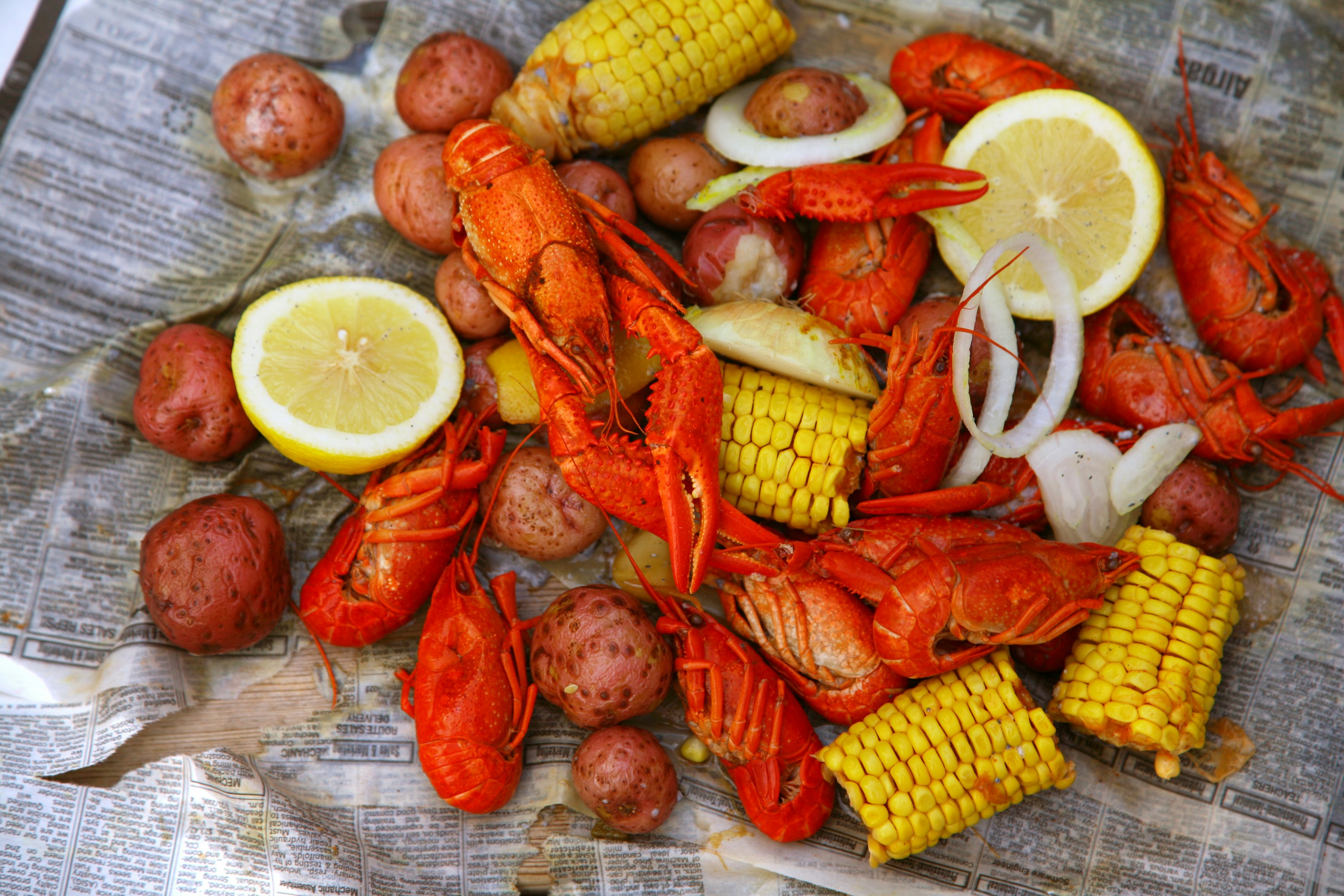 A pile of crawfish, potatoes, corn on the cob with a side of onions and a lemon wedge lay on old newspaper.