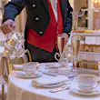 A smartly dressed waiter in a red waistcoat, seen at the mid-section only, pours tea from a shiny silver jug. A two-tier tray of cakes stands nearby, along with glasses of bubbly