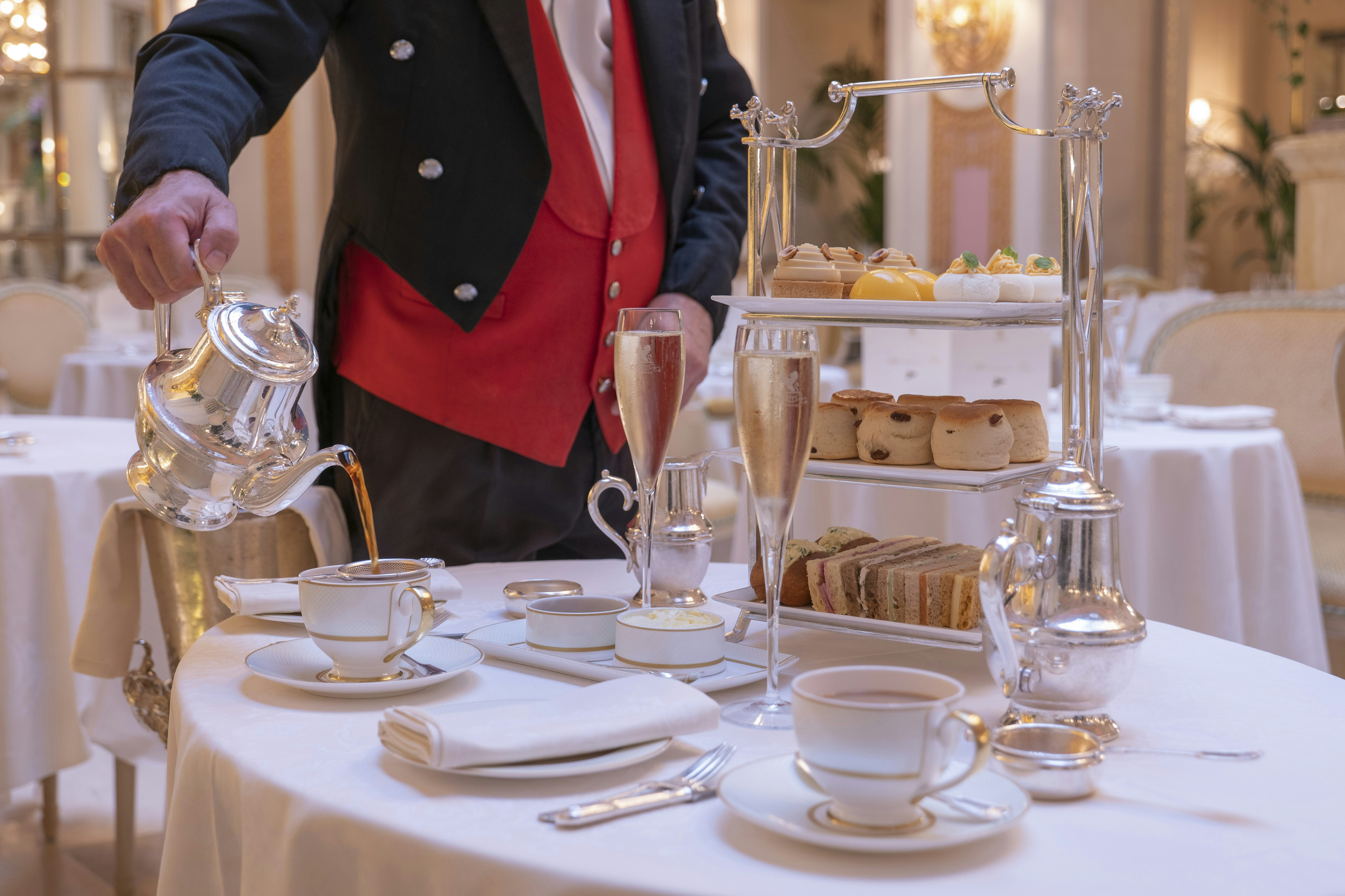 A smartly dressed waiter in a red waistcoat, seen at the mid-section only, pours tea from a shiny silver jug. A two-tier tray of cakes stands nearby, along with glasses of bubbly
