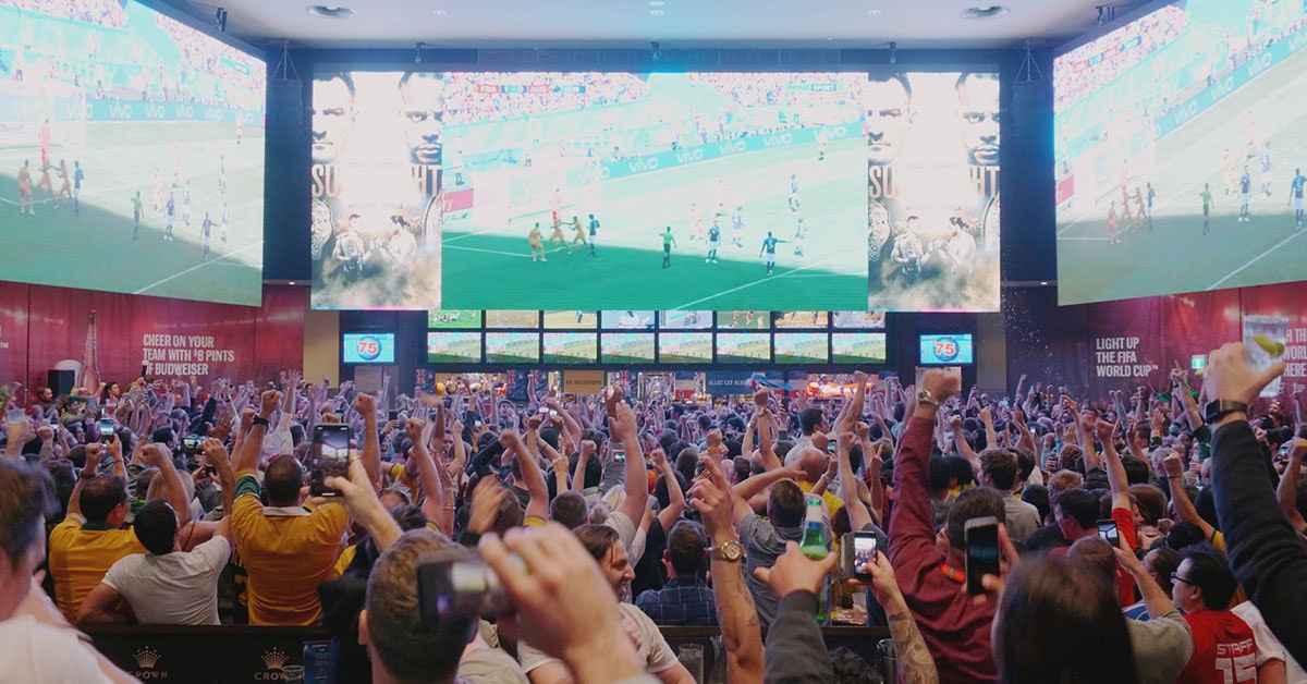 A huge sports bar, with enormous screens showing a football match. The room is packed with people raising their hands and cheering.