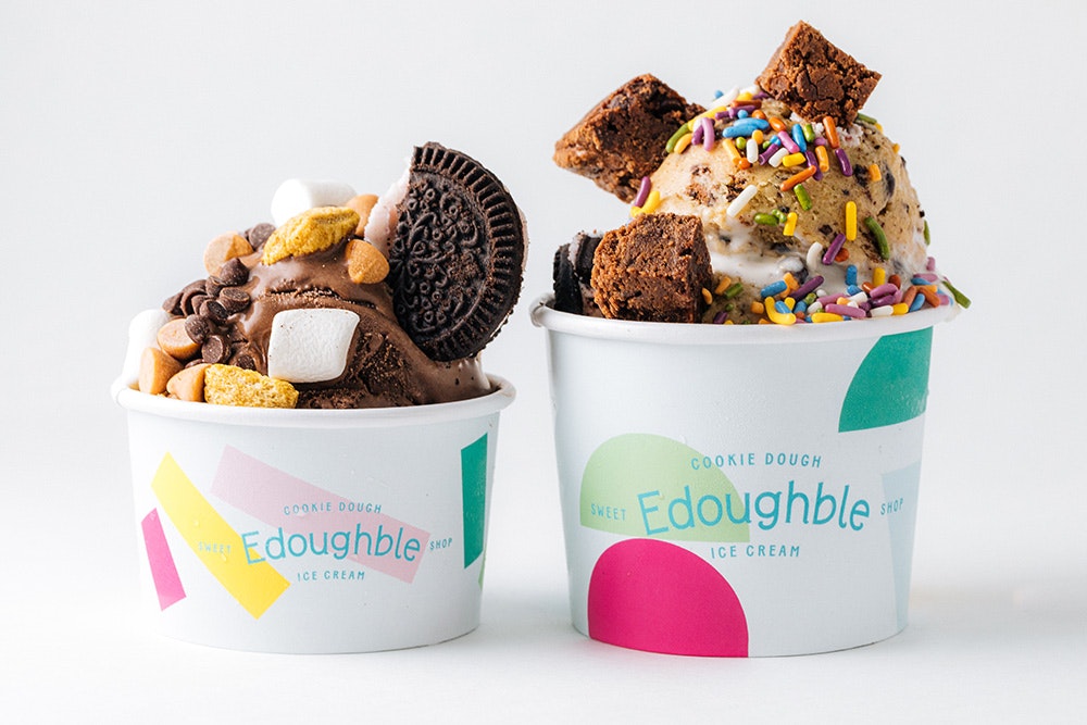 Two Edoughble cups holding cookie dough with cookies, marshmallows, sprinkles, and other mix-ins