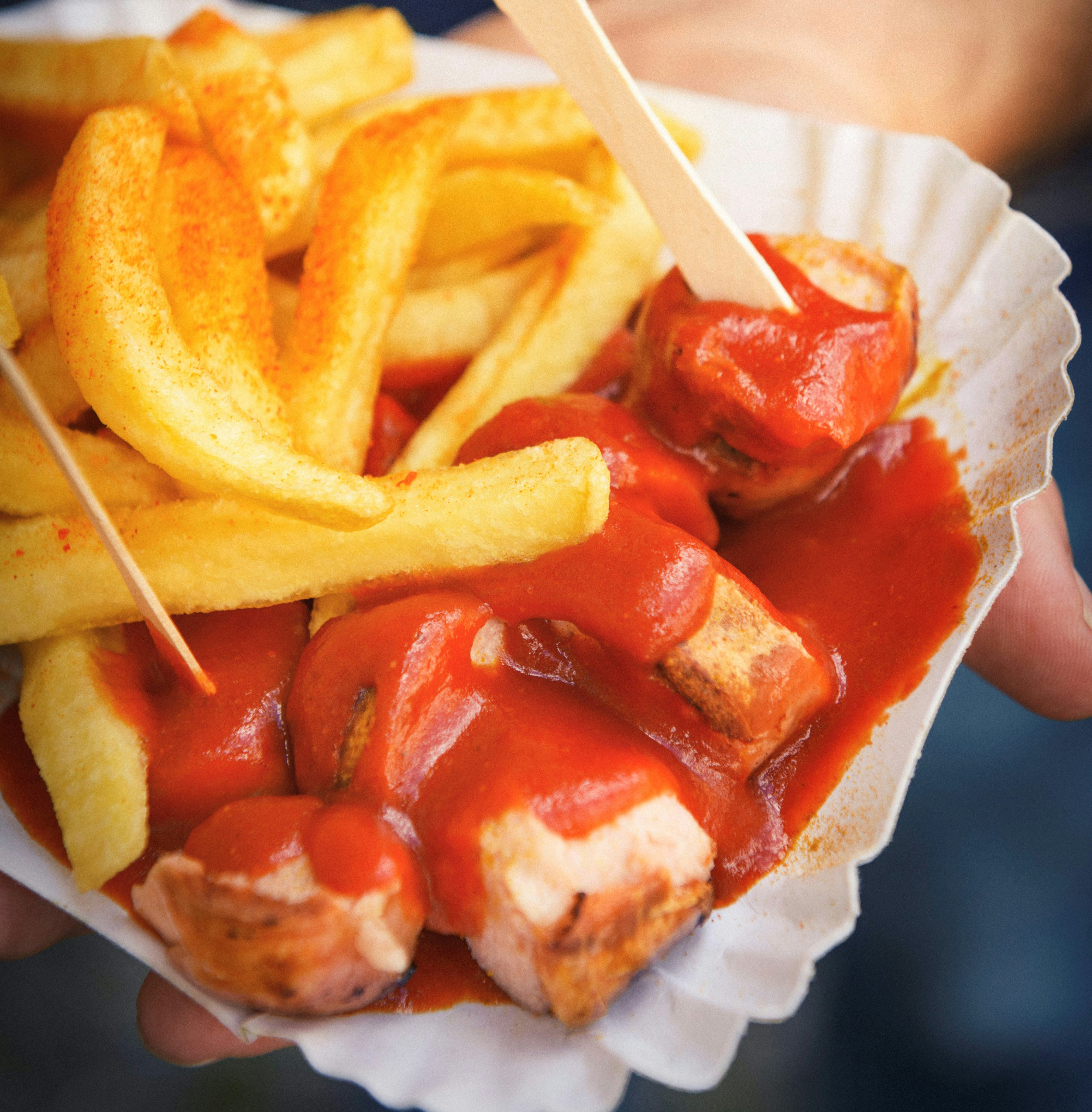 A hand holds out a portion of currywurst, purchased on a night out in Berlin. The food, cut up sausage and chips, is slathered in curry sauce.