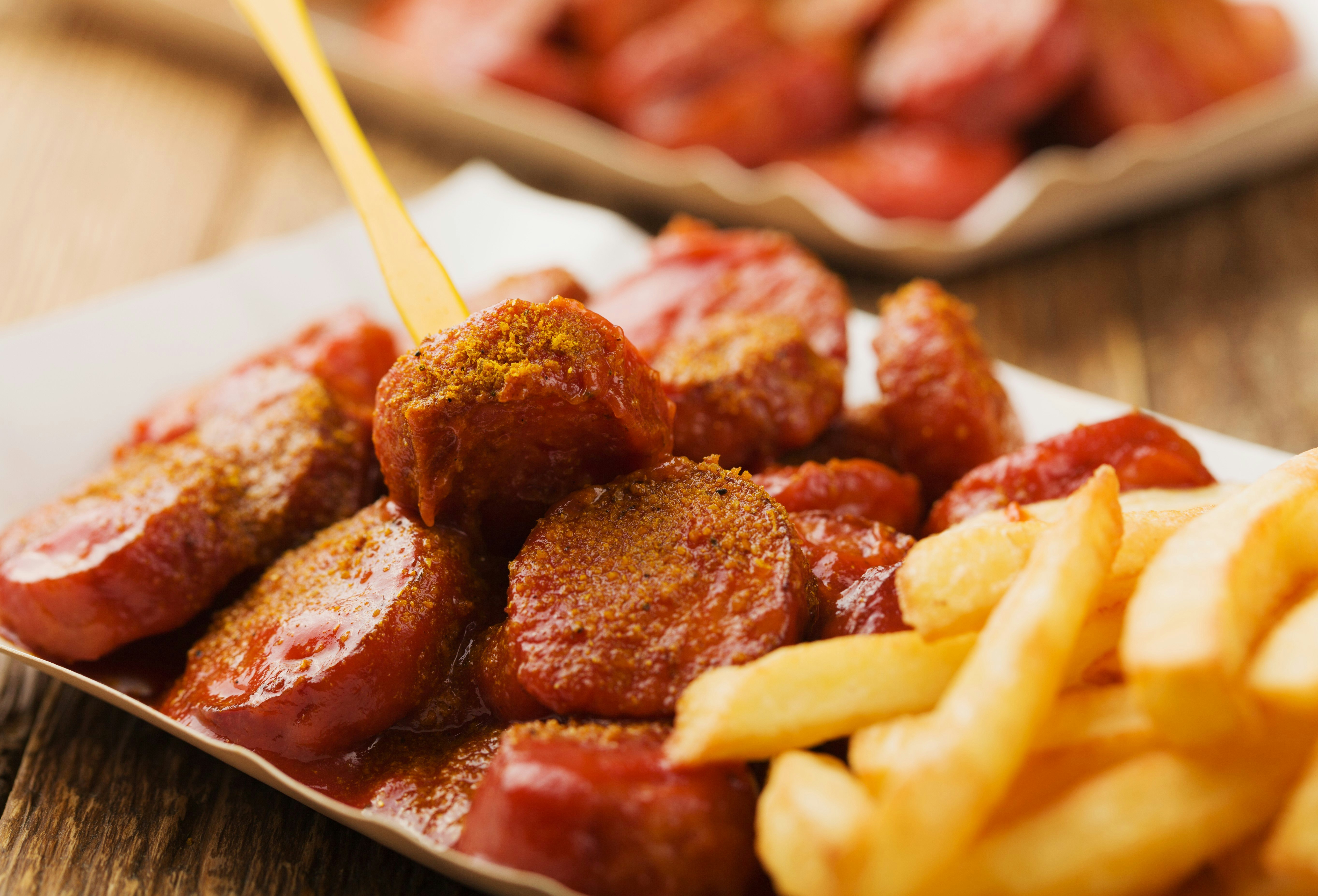 A close-up view of currywurst (cut up sausage with curry sauce) served with chips on a paper plate.