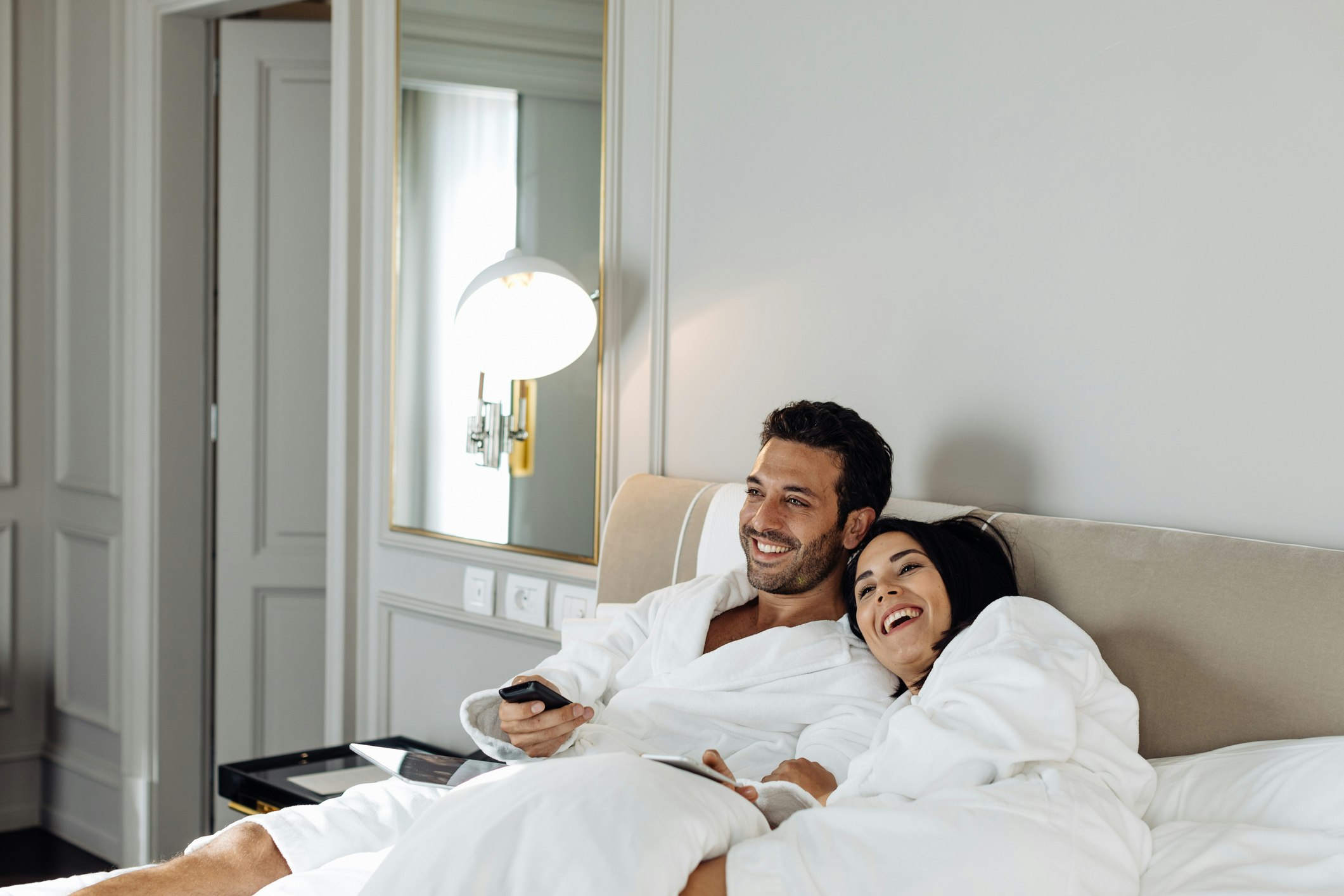 Couple in white bathrobes sit on a bed in a hotel suite