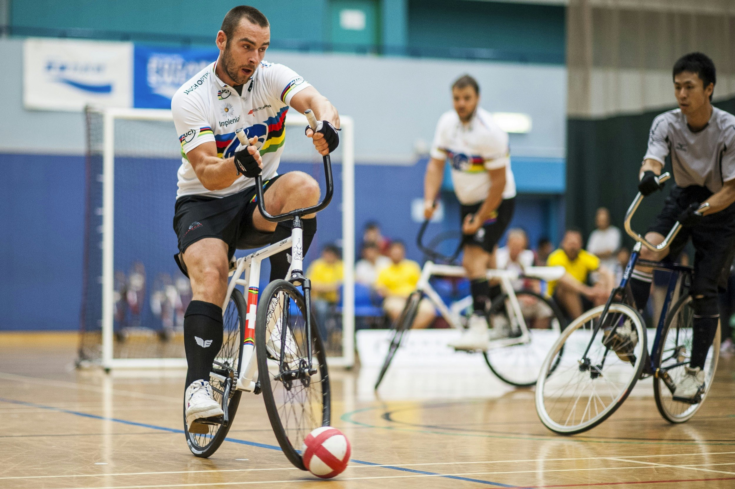 A man uses a specialty bicycle to hit a small ball across a hard gym floor; unique sporting events