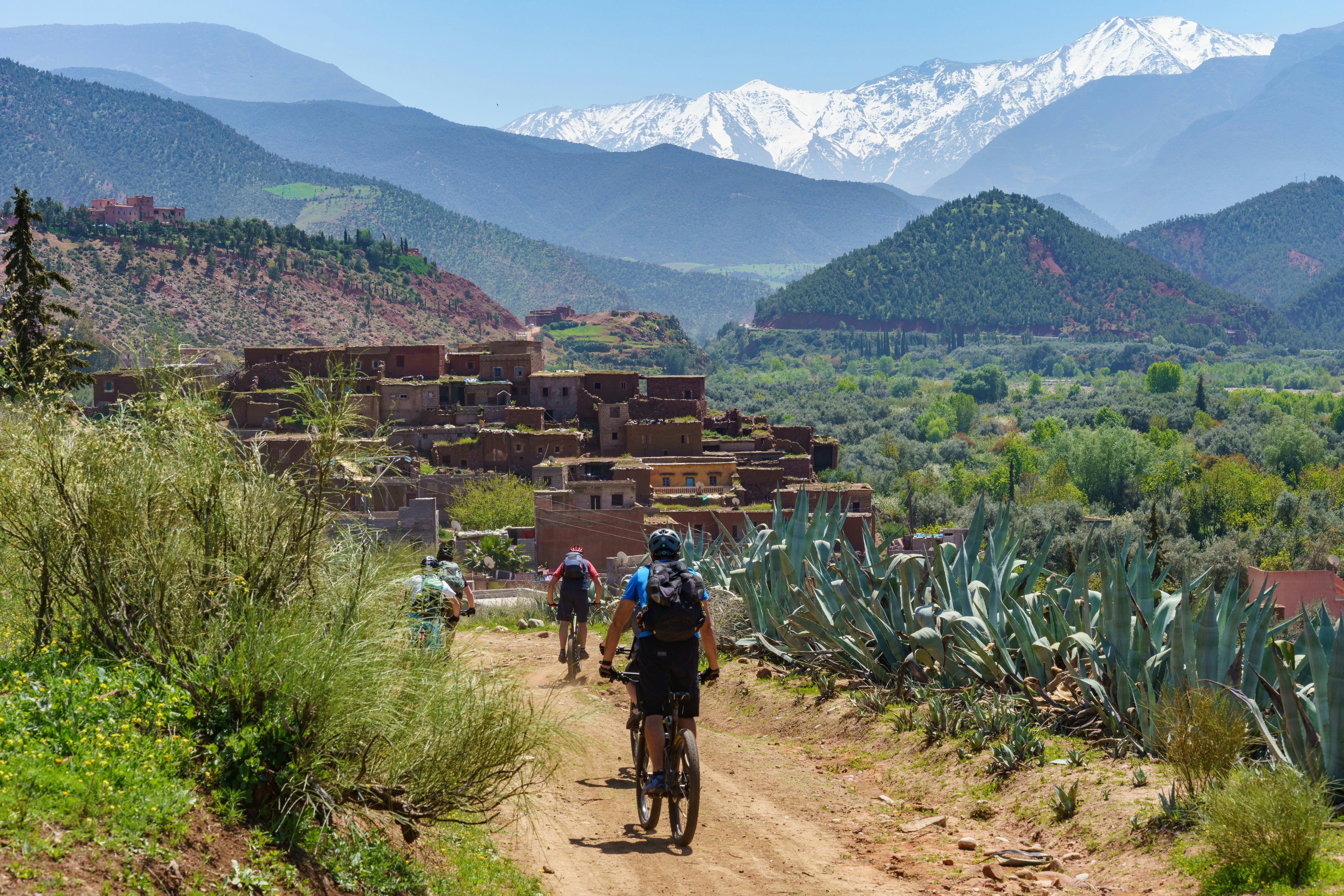 A group of cyclists pedal along a trail towards a traditional-looking village in the hills of the Atlas Mountains, Morocco.
